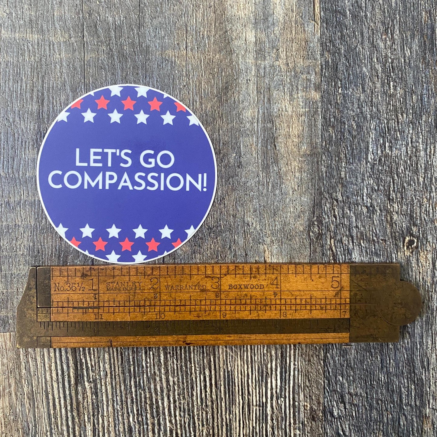 Sticker with 'LET'S GO COMPASSION!' text on a wooden ruler against a wooden background.