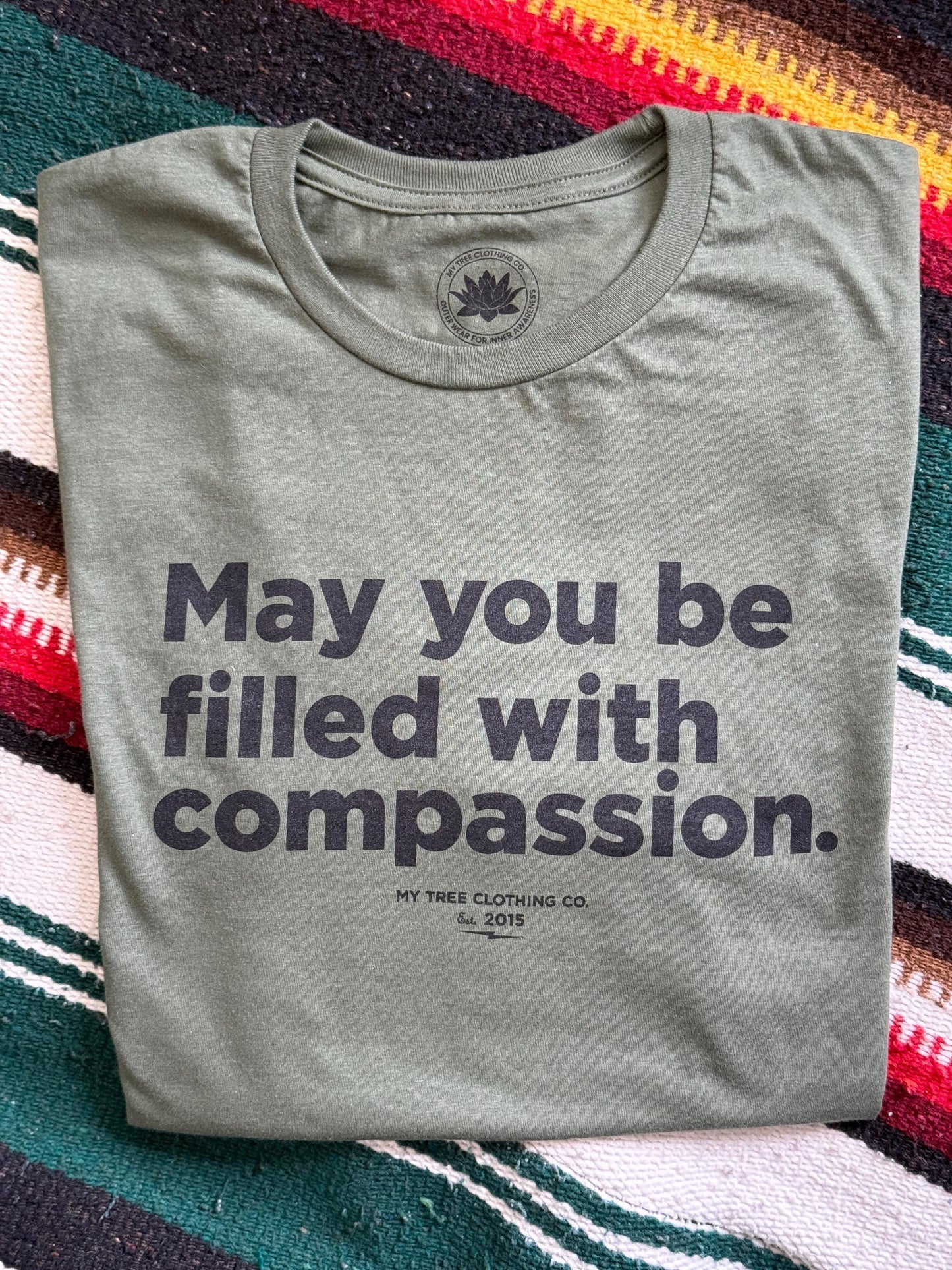 Folded gray t-shirt with text, may you be filled with compassion, on a colorful striped fabric background