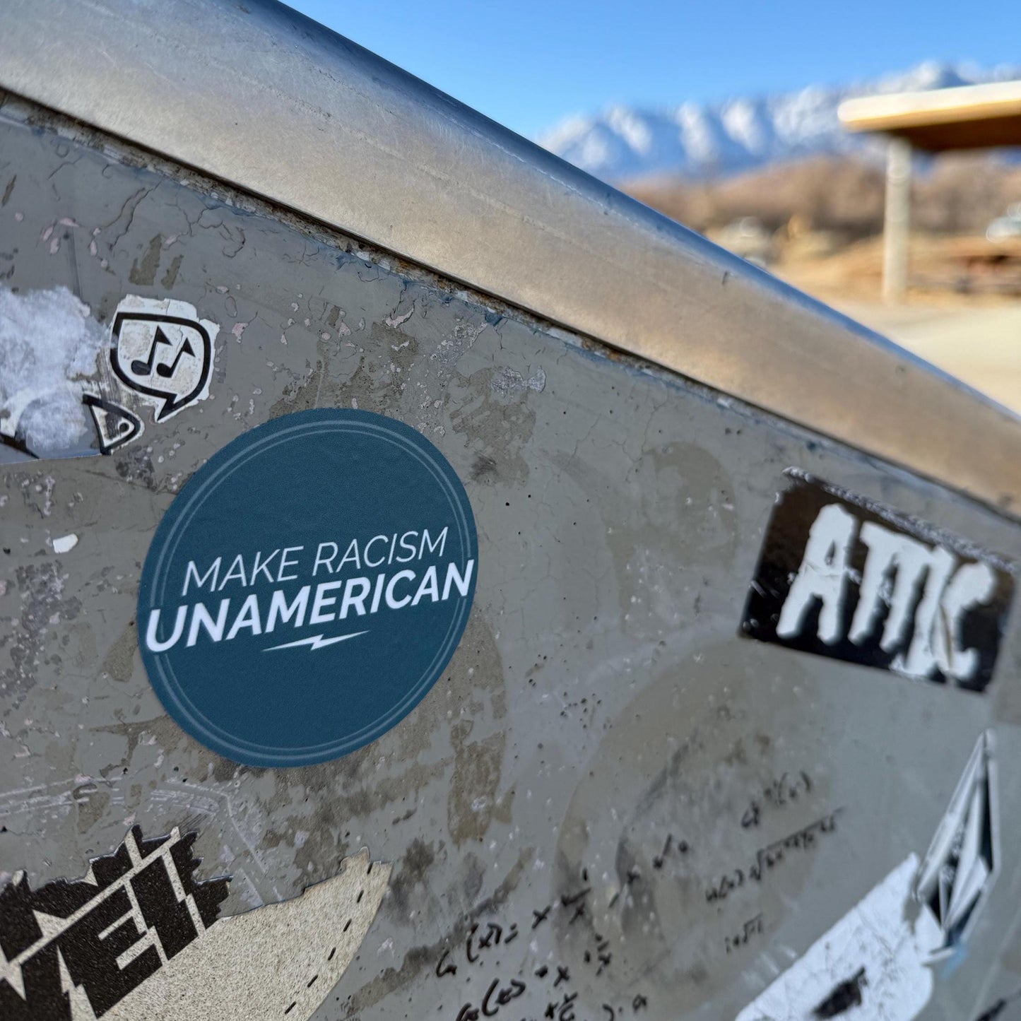 2.5" 'Make Racism UNAMERICAN' Sticker on a metal surface with mountains in the background.