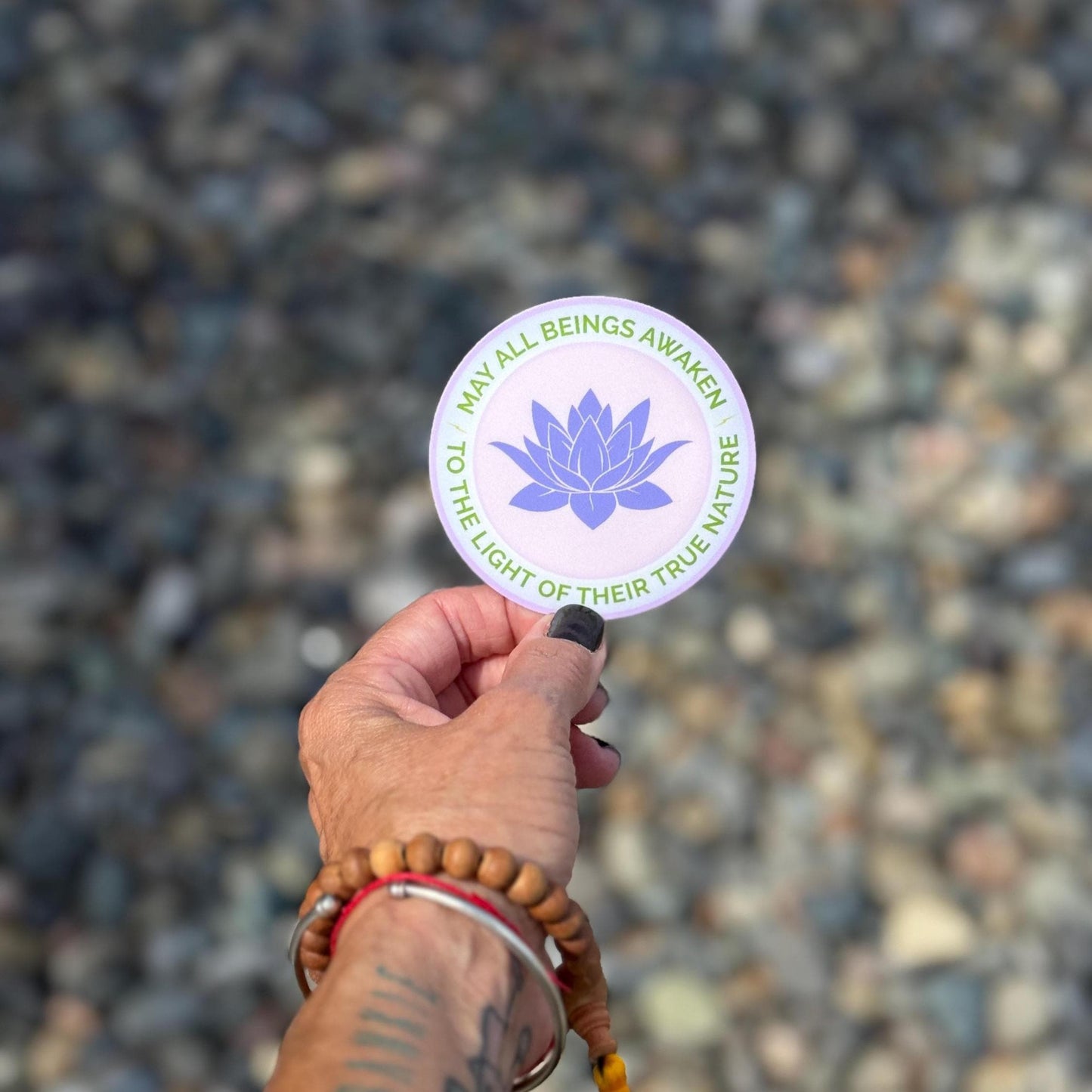 Hand holding a 3" round sticker with a purple lotus flower design and the text, may all beings awaken to the light of their true nature, against a blurred natural background.
