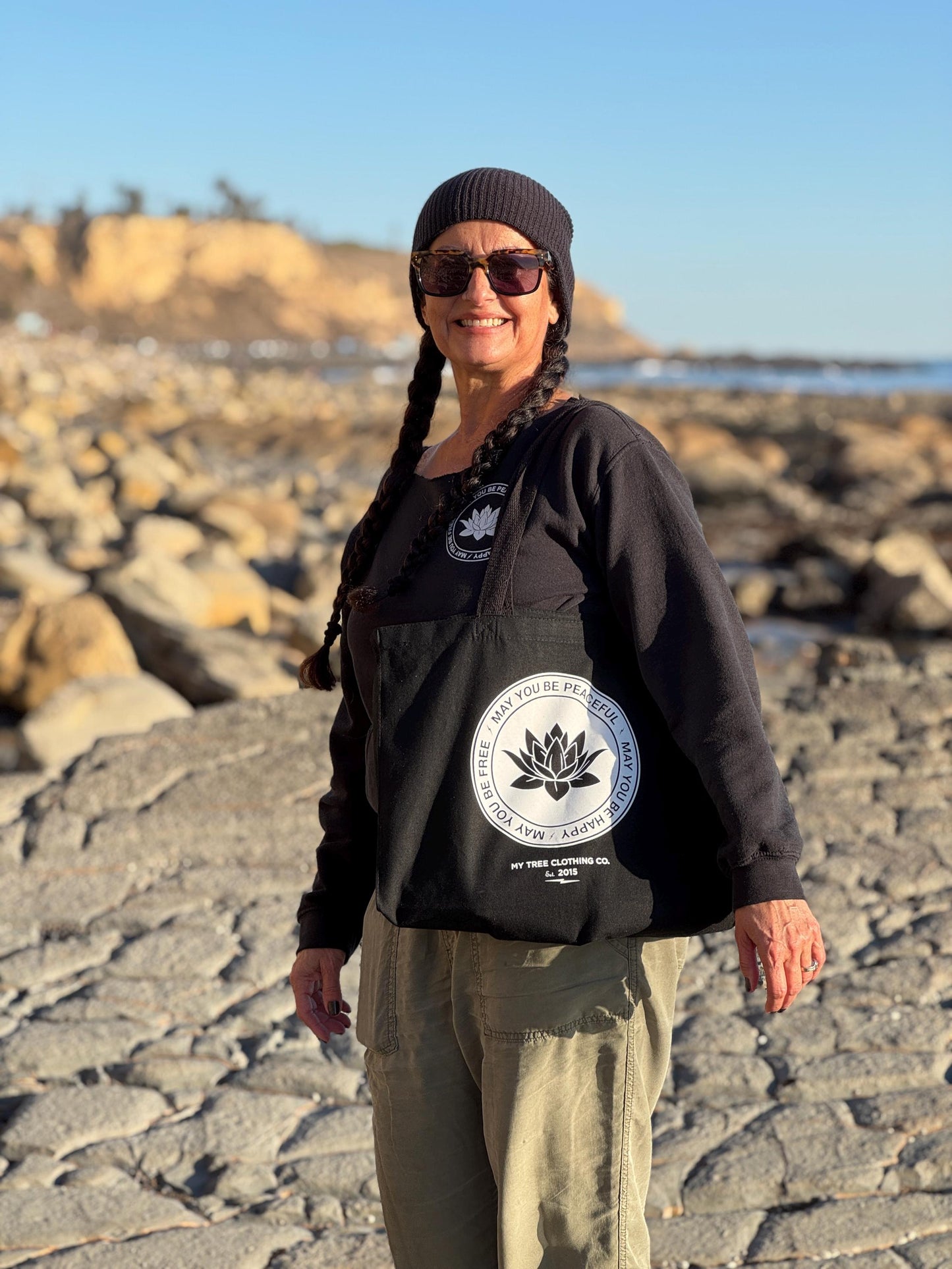 Person wearing a black hoodie with a logo on a rocky beach holding a black tote bag with a lotus and the text, may you be happy may you be peaceful may you be free.