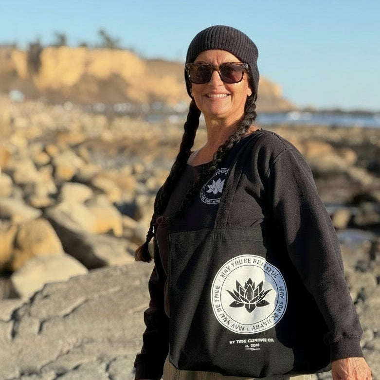 Person wearing a black hoodie with a logo on a rocky beach holding a black tote bag with a lotus and the text, may you be happy may you be peaceful may you be free.