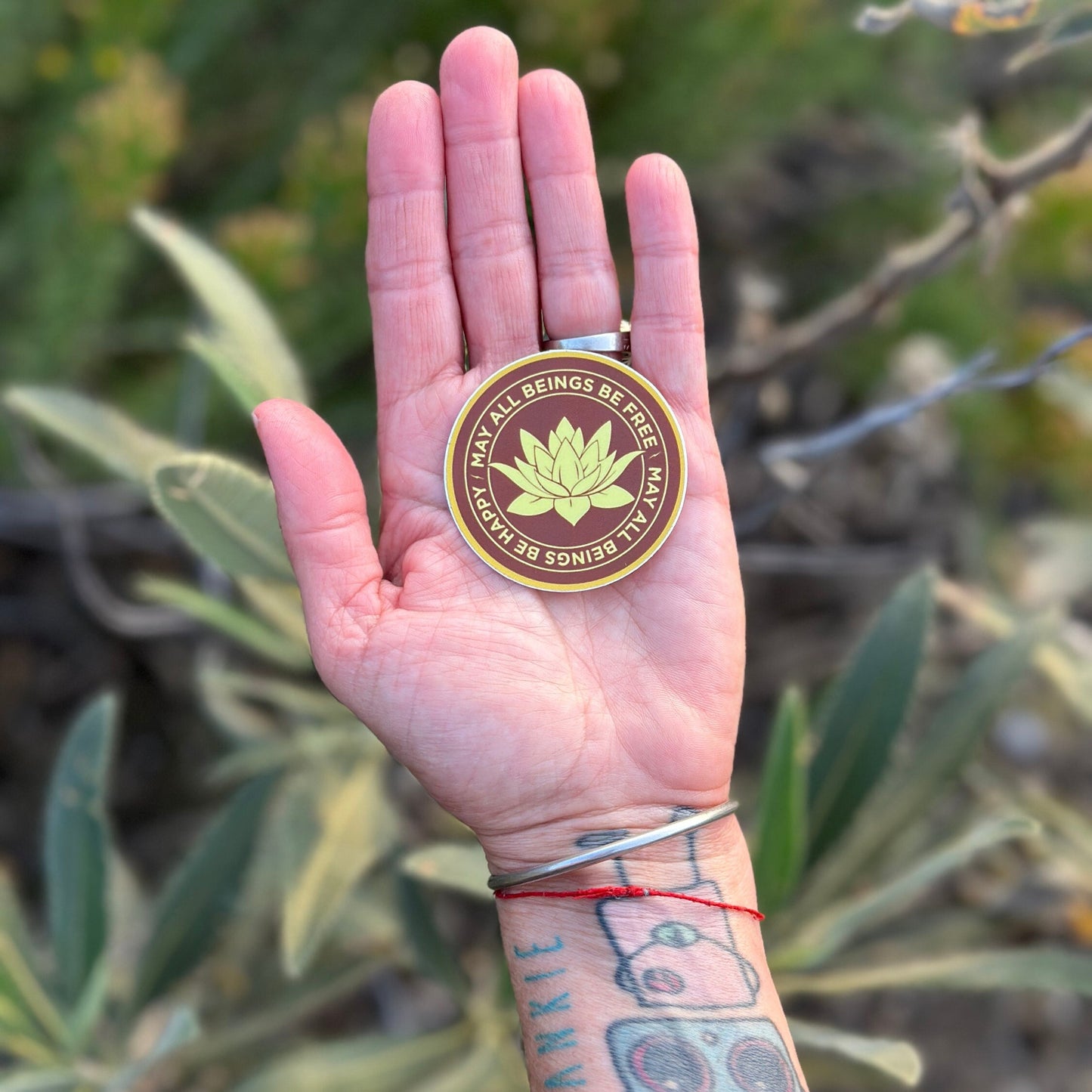 Hand holding a 2" round saffron and gold vinyl sticker with Buddhist mettā prayer text, may all beings be free, may all being be happy, against a natural background.