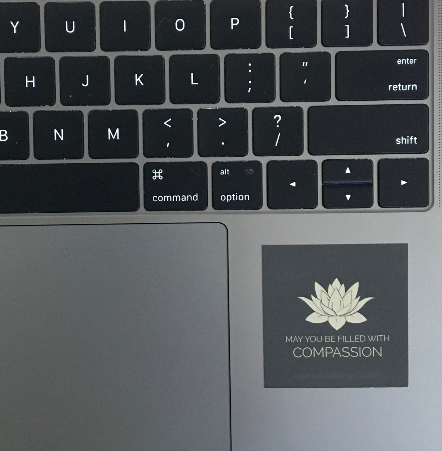 Laptop keyboard with a 2" square lotus flower sticker with text 'May you be filled with compassion' sticker on the palm rest area.