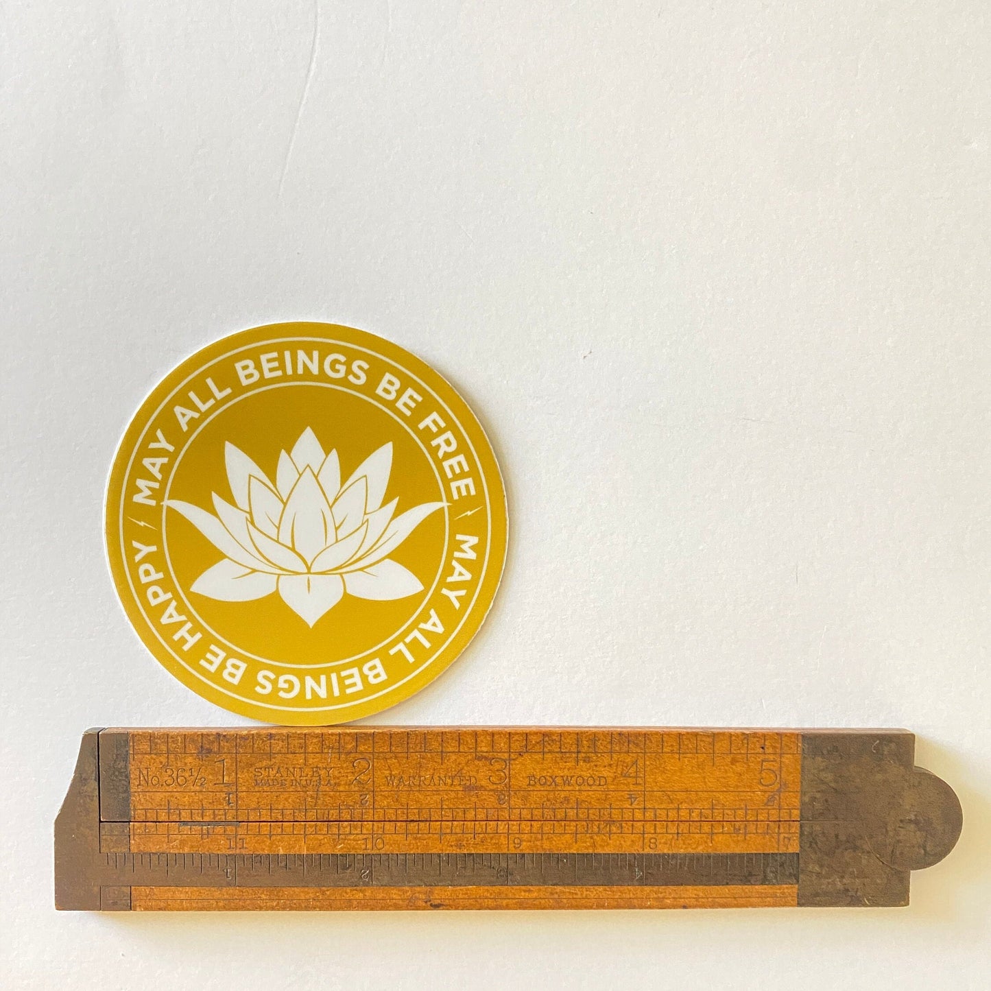 Yellow circular sticker with a white lotus flower design and text, placed on a white background with a wooden ruler below.