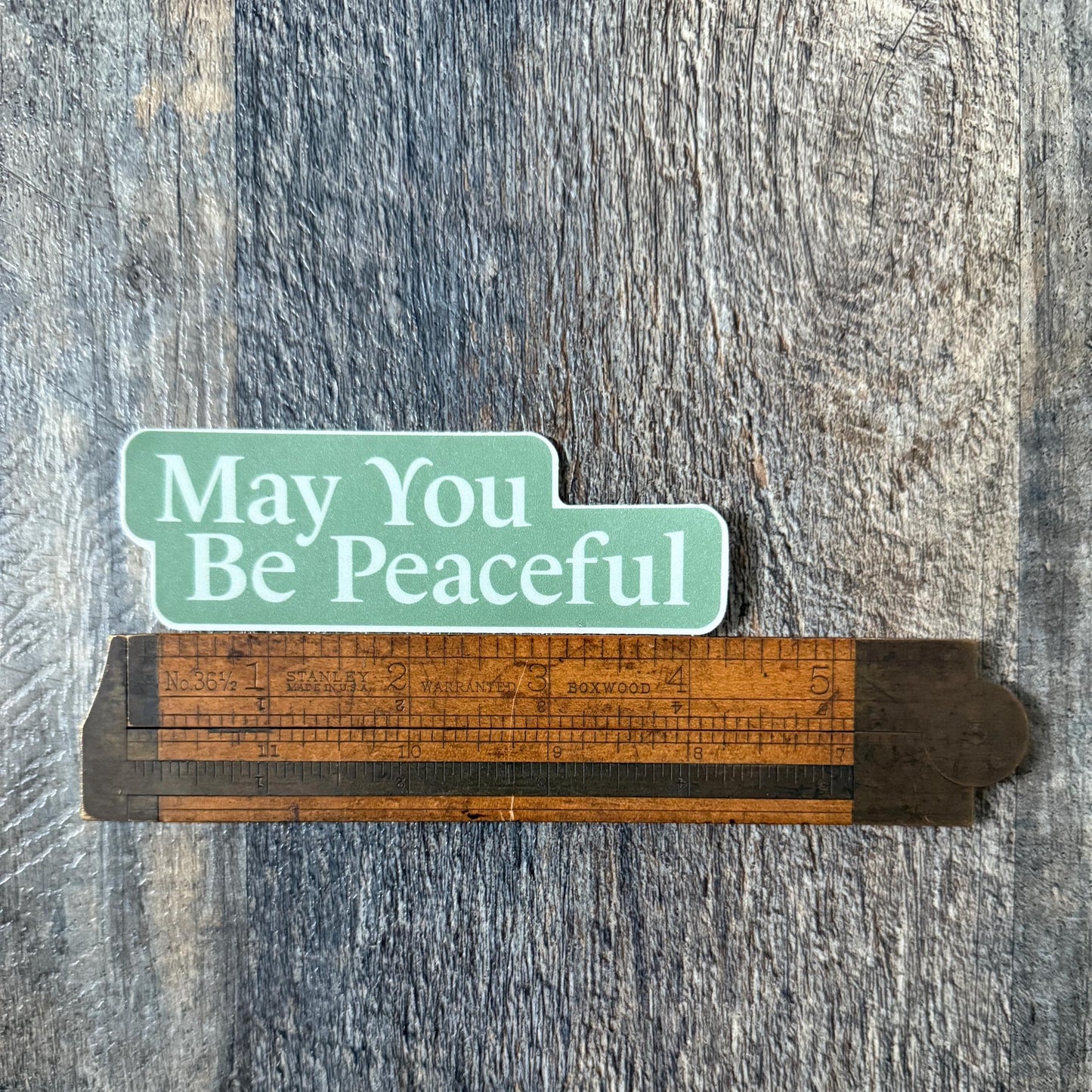 Wooden ruler with 'May You Be Peaceful' sticker on a textured surface.