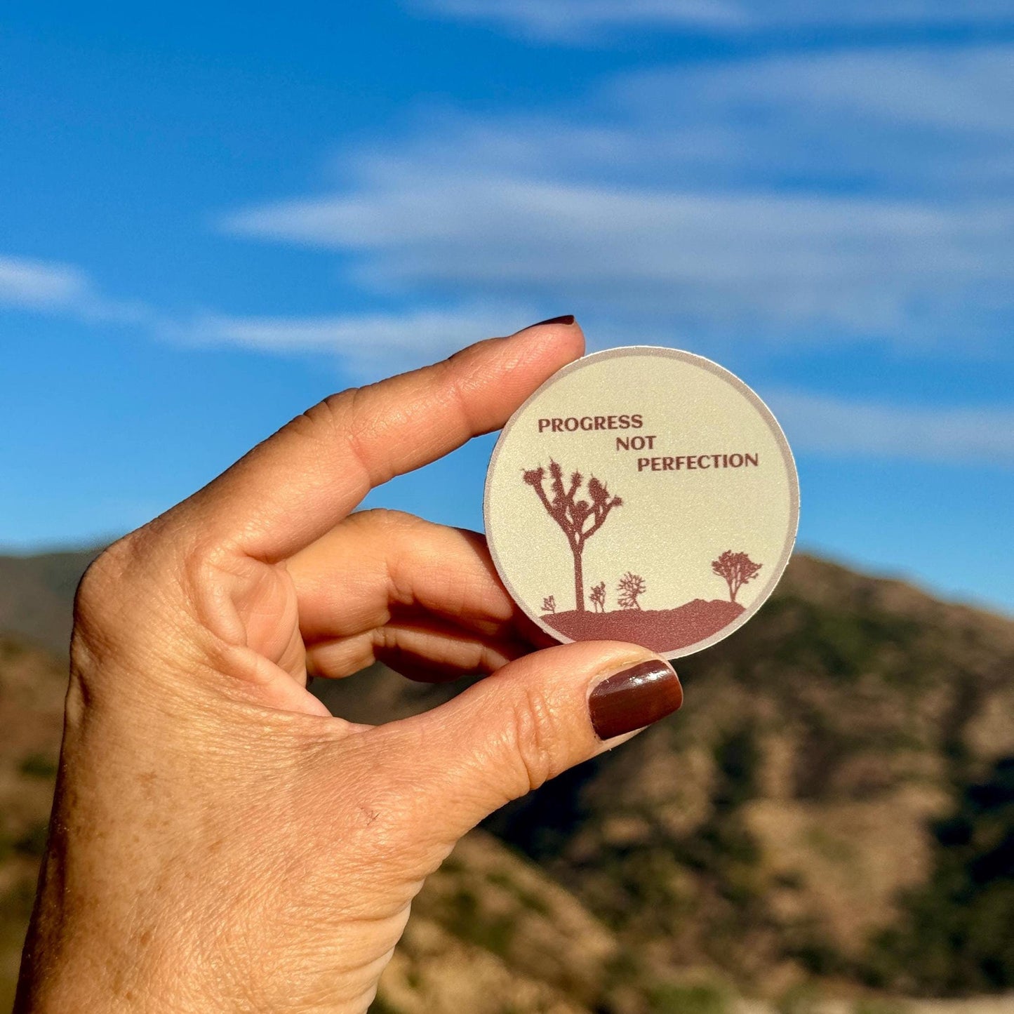 Hand holding a circular sticker with desert landscape design and 'Progress Not Perfection' text against a mountainous background.