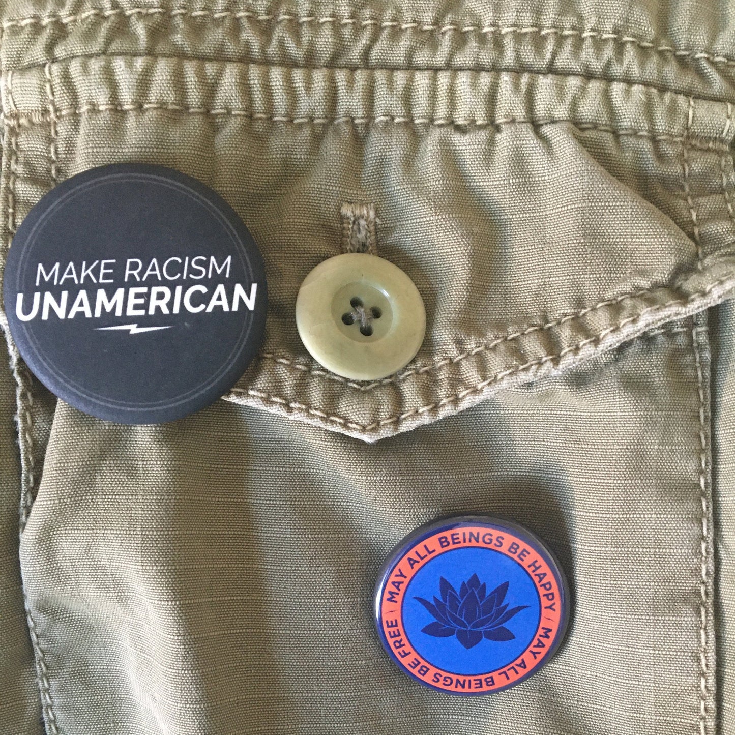Two buttons on a jacket: one with 'Make Racism Unamerican' text and the other is a 1.25" circular pin back button with a lotus flower design and text, may all beings be free may all beings be happy.