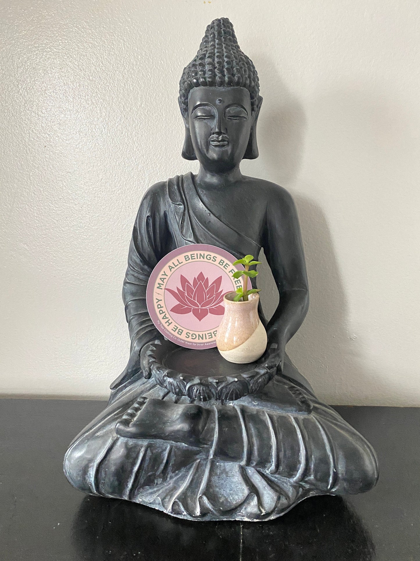 Black Buddha statue holding a plant with a 4" Circular sticker with lotus flower design and the text, MAY ALL BEINGS BE FREE MAY ALL BEINGS BE HAPPY.