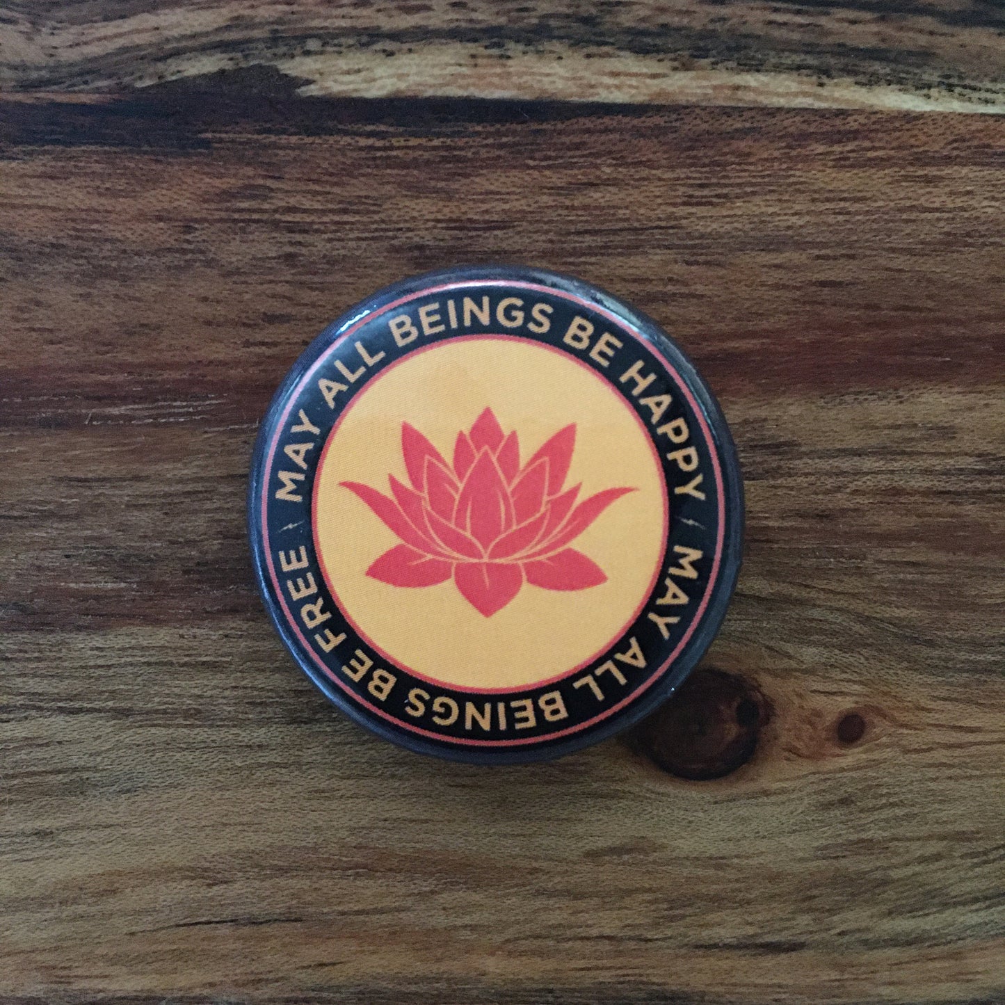 1.25" Orange round pin back button with a buddhist lotus flower and the text 'may all beings be happy may all beings be free,' on a wooden surface.