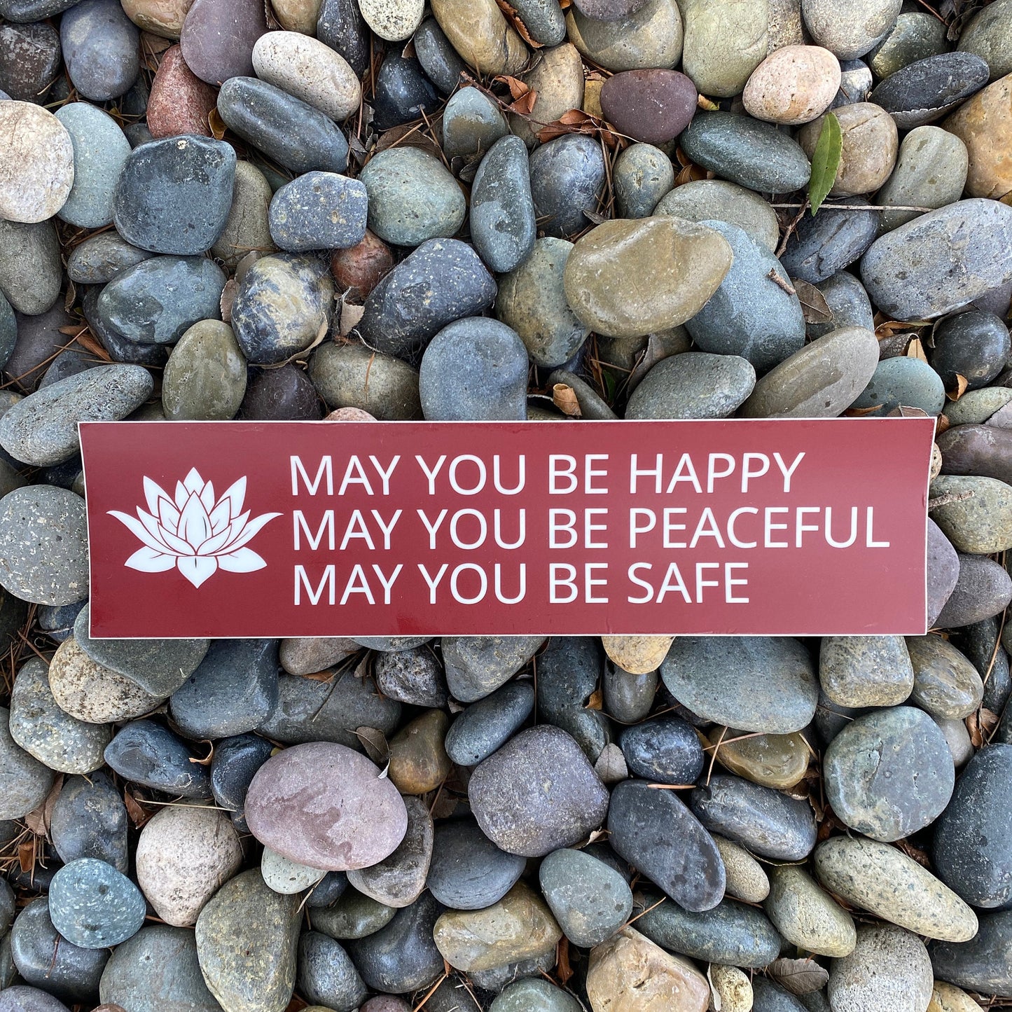 Red and white bumper sticker with a lotus and the prayer, may you be happy may you be peaceful may you be safe, on a bed of multicolored stones.