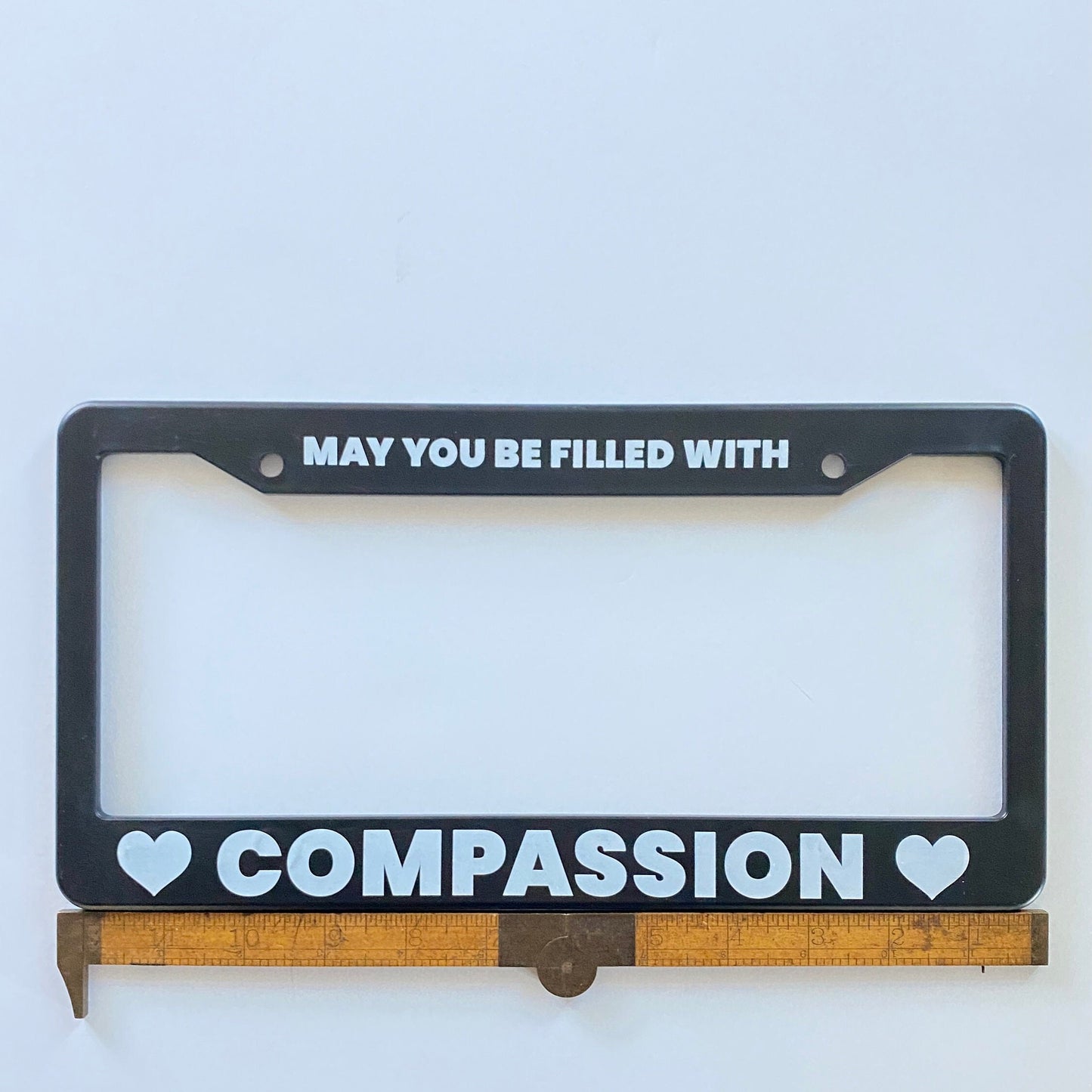 Black license plate frame with text 'MAY YOU BE FILLED WITH COMPASSION' on a white background, being measured by an antique ruler.