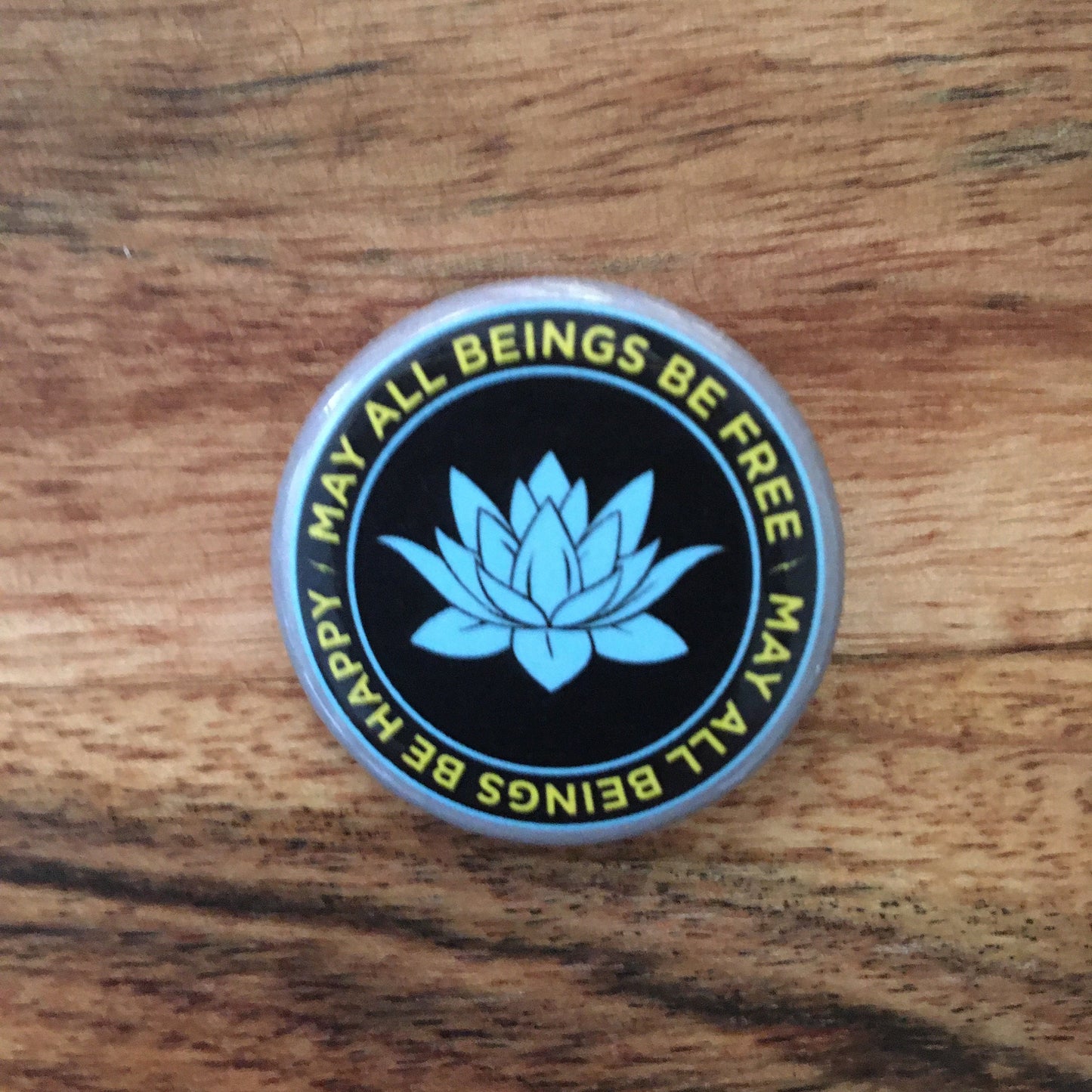 1.25" black and blue lotus button with the Buddhist prayer, may all beings be free may all beings be happy, on a wooden surface.
