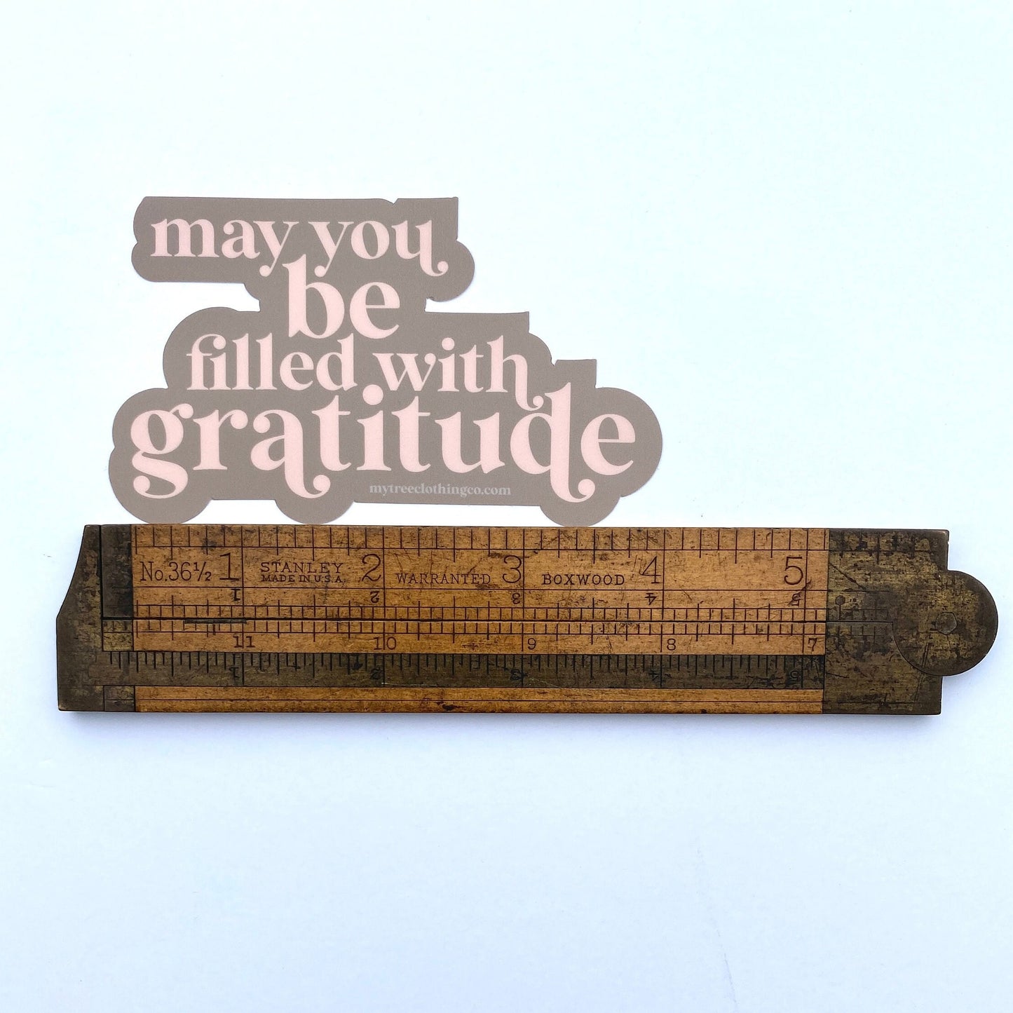 Decorative wooden ruler with a pink and brown 'may you be filled with gratitude' sticker on a light blue background.