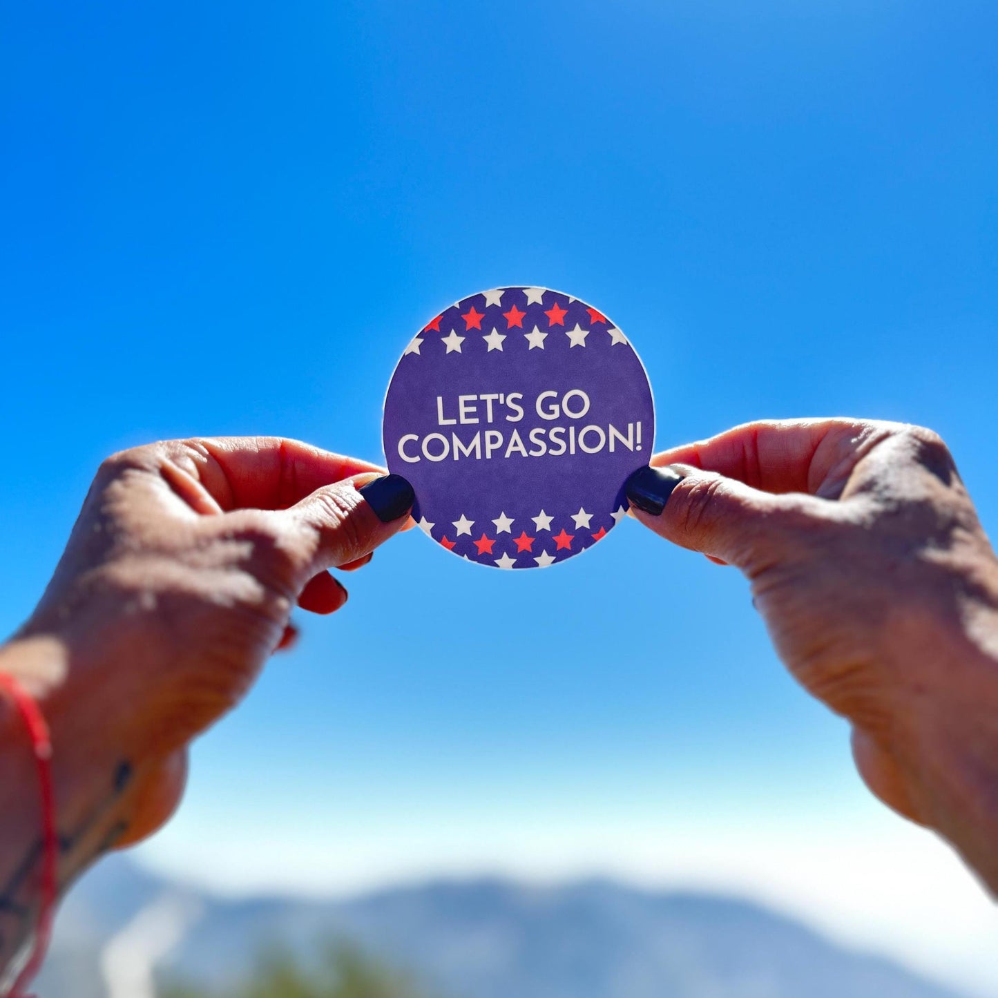 Two hands holding a sticker with patriotic red, white, and blue, 'Let's Go Compassion!' sticker with a clear blue sky in the background. Real patriots support kindness, inclusion, and diversity!