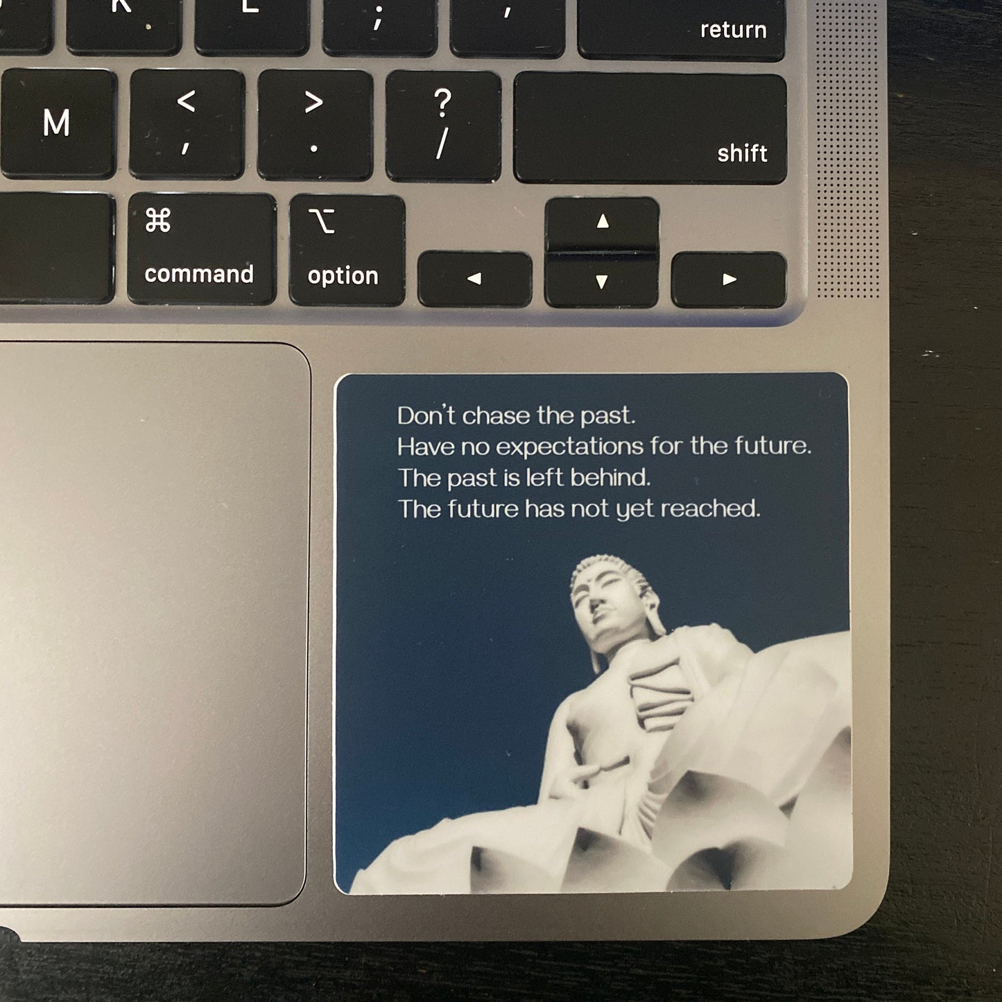 Laptop with a 3" round sticker with a Buddha statue and text, "don't chase the past, have no expectations for the future, the past is left behind, the future has not yet reached" on the palm rest area.