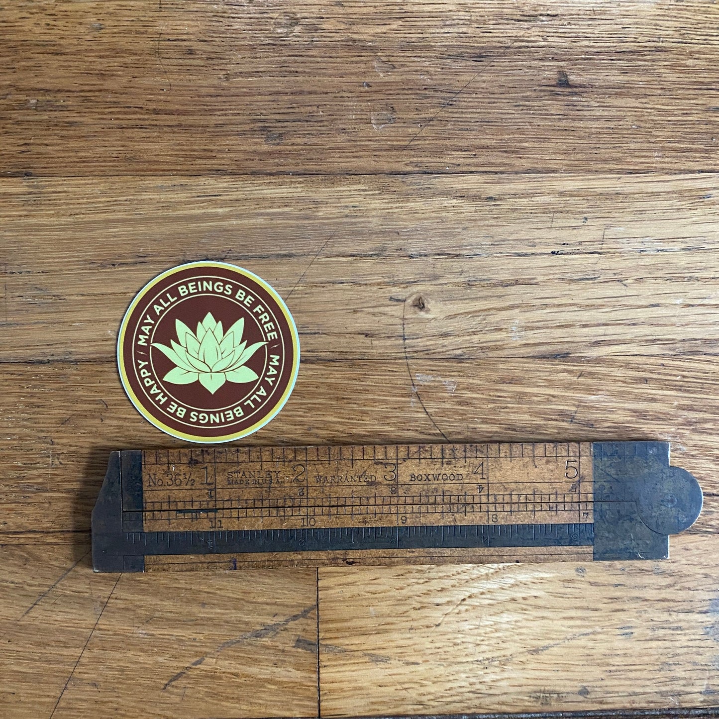2" round saffron and gold vinyl sticker with a Buddhist mettā prayer text, may all beings be free, may all being be happy, on a wooden surface with a ruler for scale.