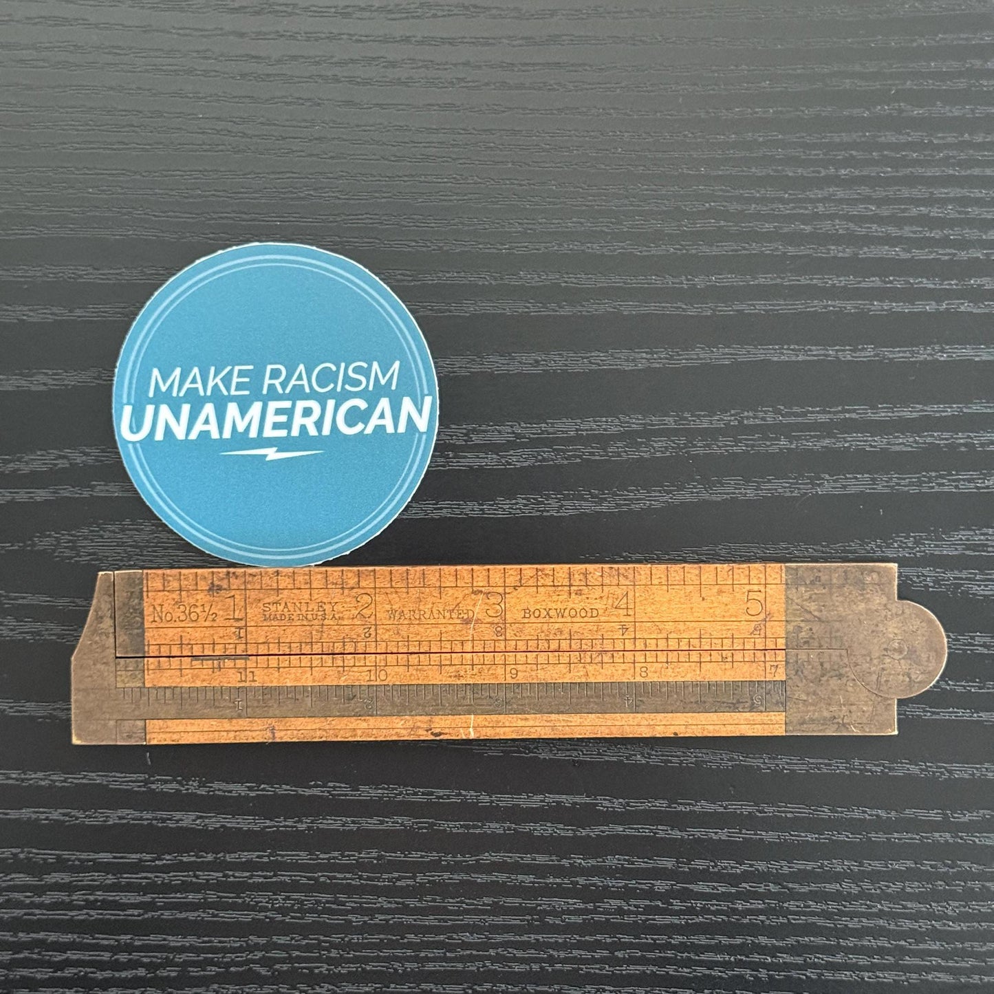 Blue circular sticker with 2.5" 'Make Racism Unamerican' text on a dark background with a wooden ruler.
