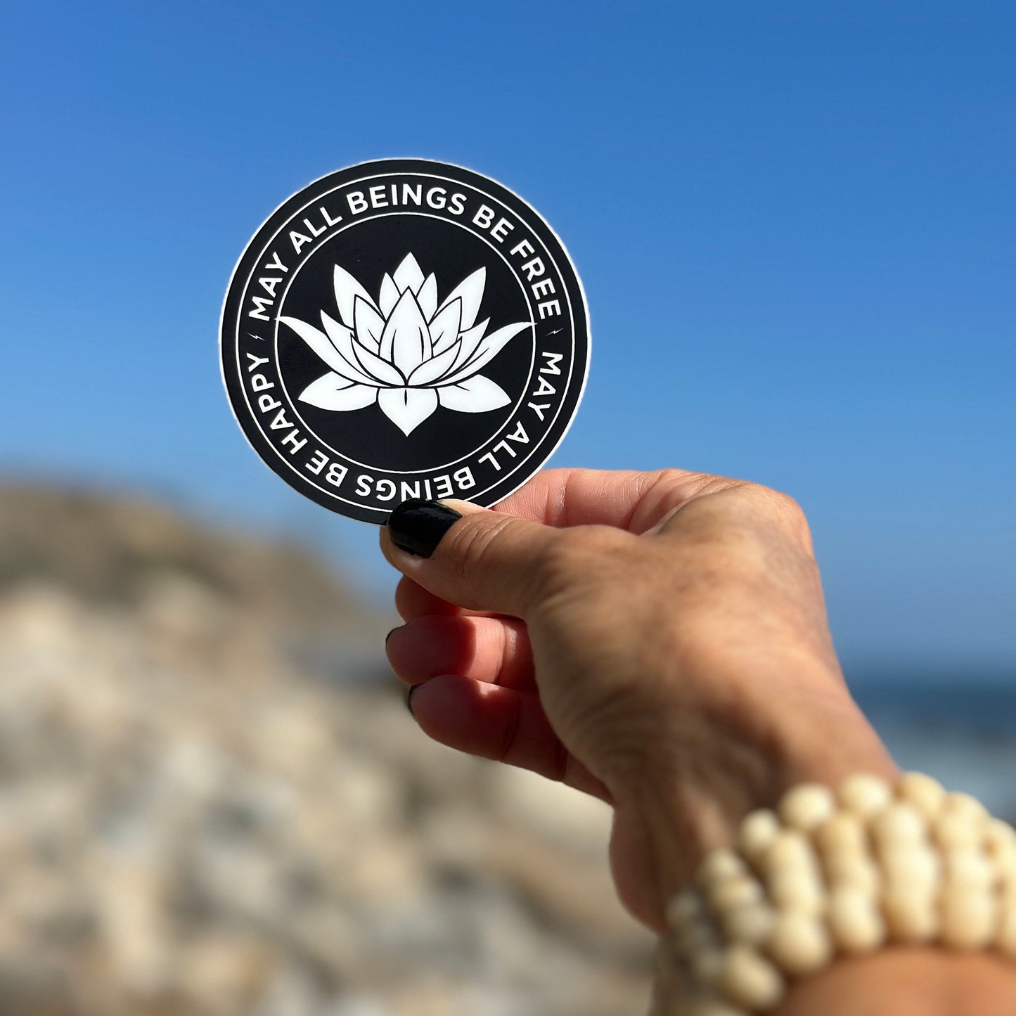Hand holding a 3" circular sticker with a white lotus flower design and text, may all beings be free may all beings be happy, against a blurred natural background