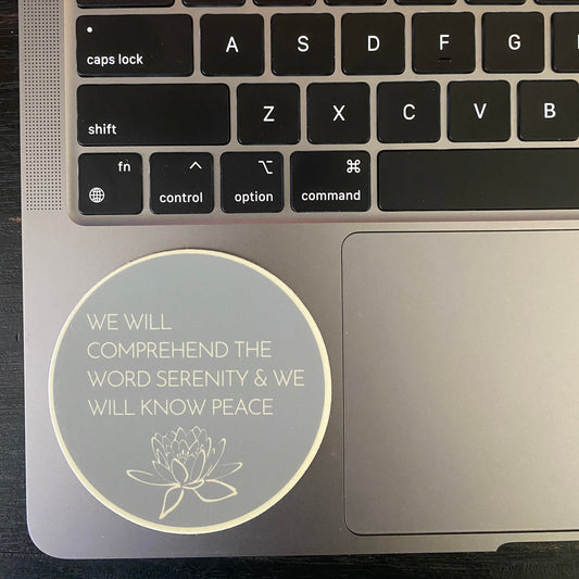 Laptop with a Round teal sticker with, WE WILL COMPREHEND THE WORD SERENITY AND WE WILL KNOW PEACE, attached to a laptop.