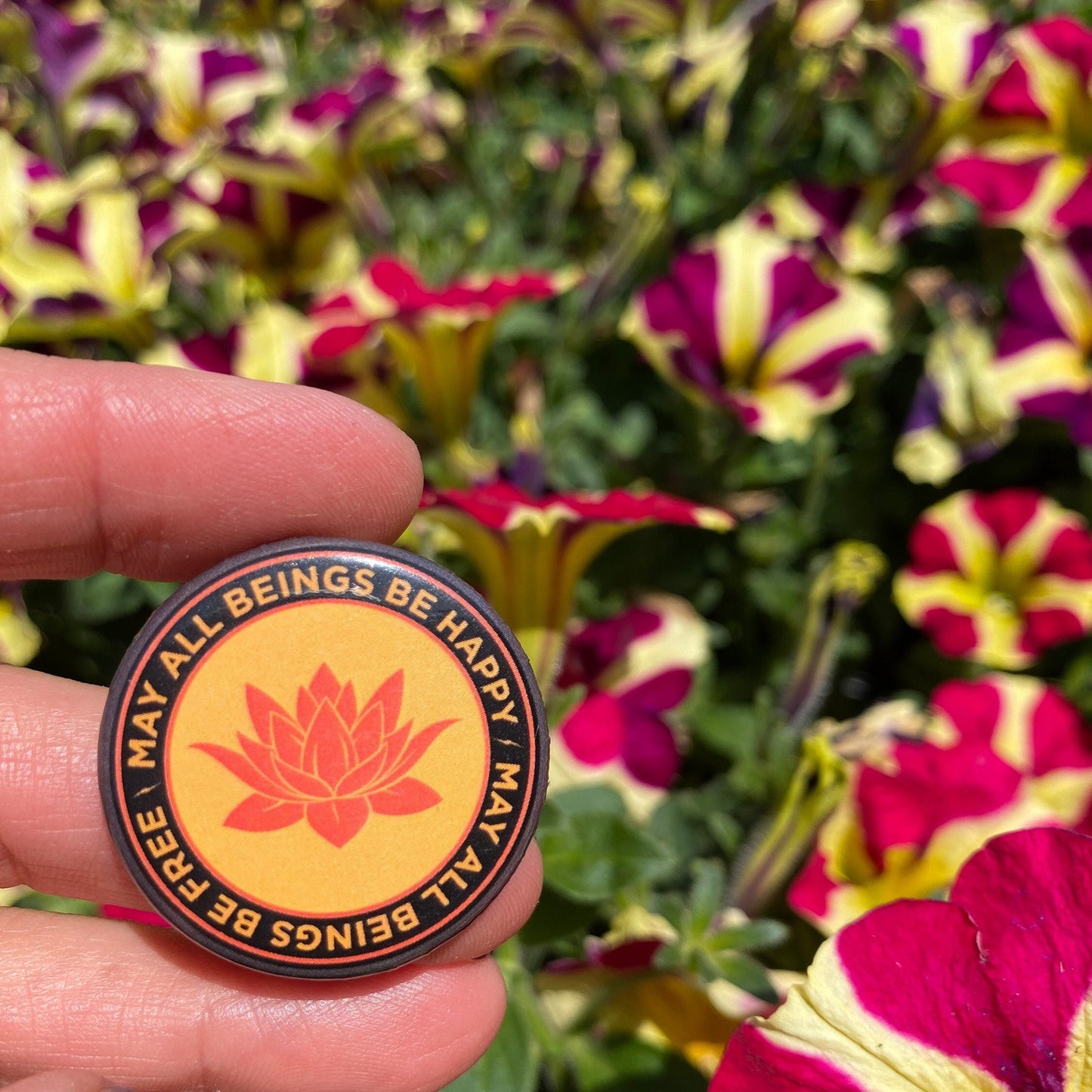 Orange round pin back button with a buddhist lotus flower and the text 'may all beings be happy may all beings be free', design held in front of colorful flowers.