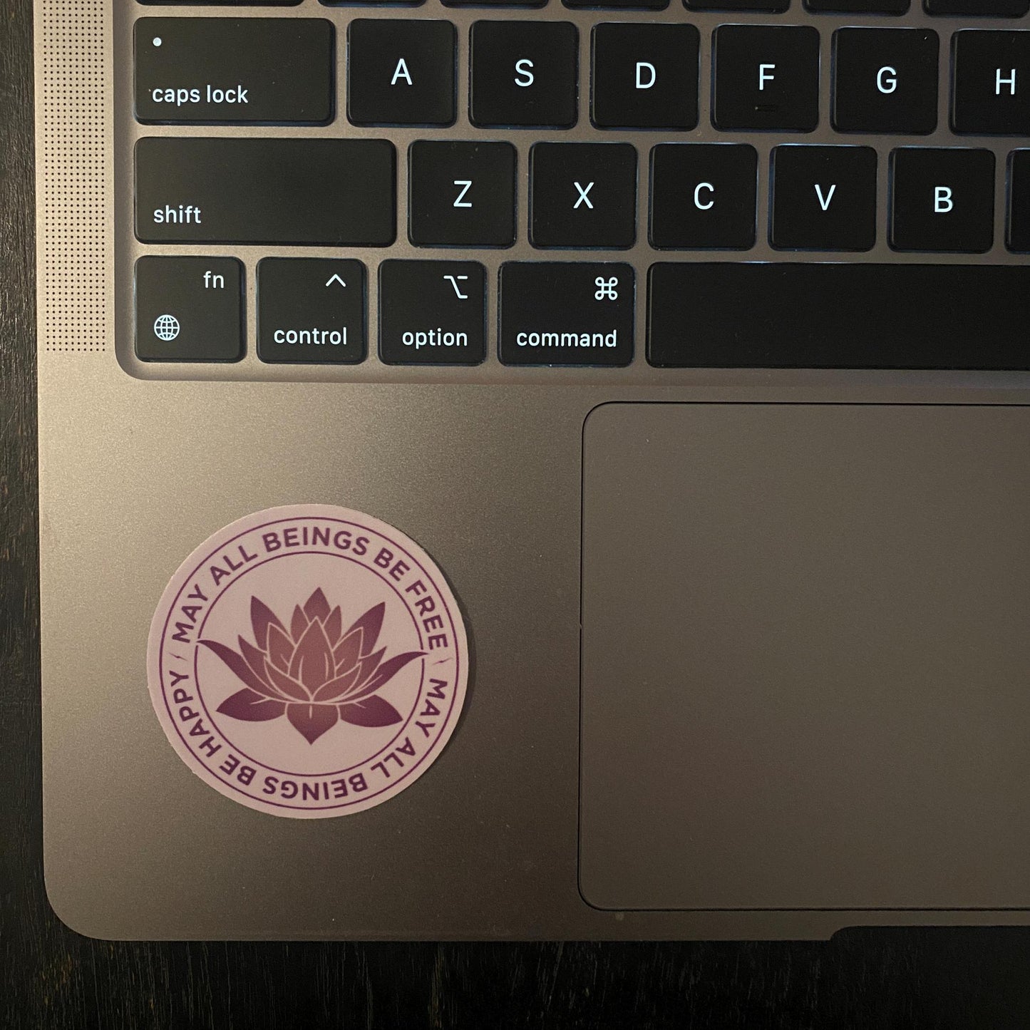 Laptop with a 2" purple sticker with a lotus design and the text, may all beings be free may all beings be happy.