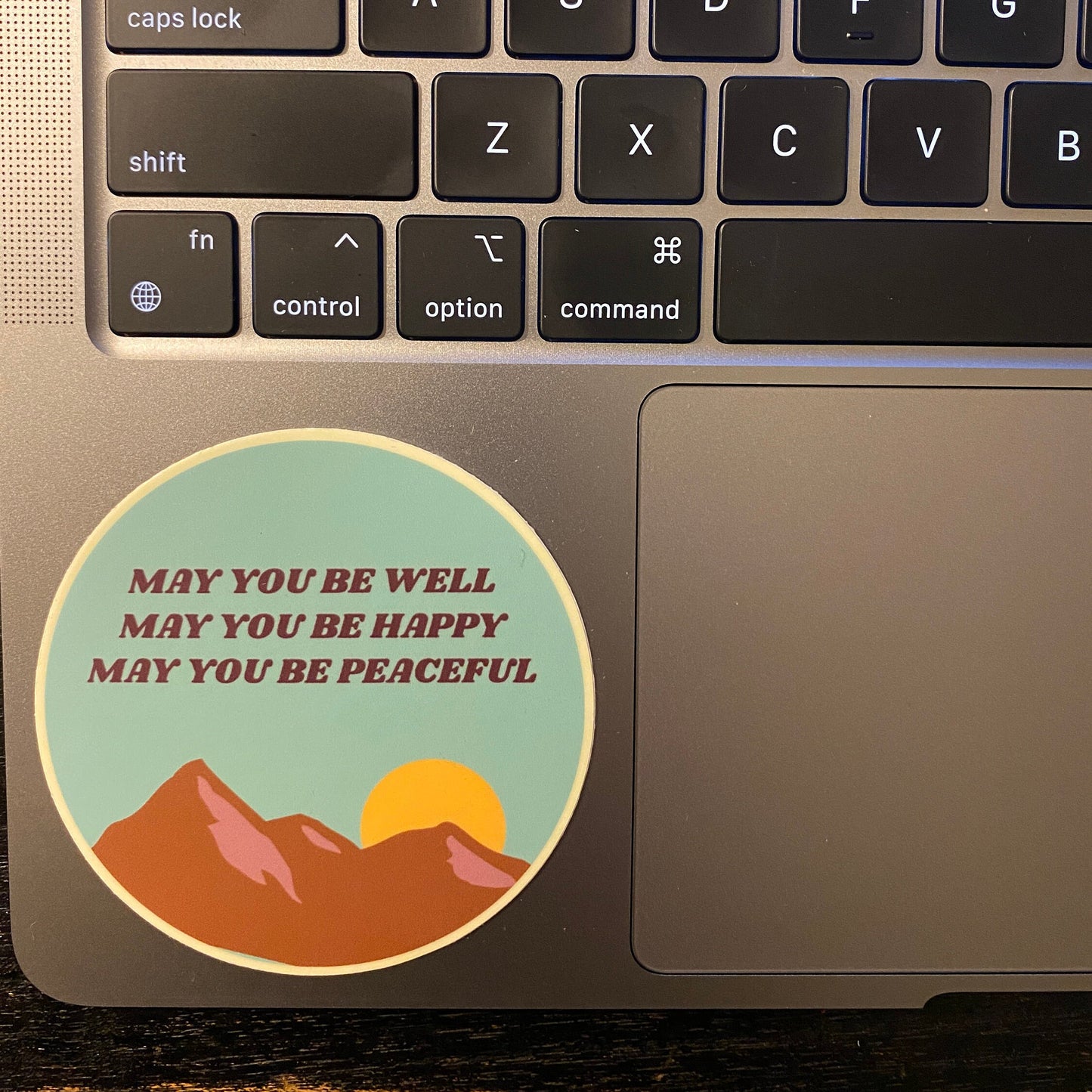 Laptop with a circular Buddhist sticker design featuring mountains and text, may you be well may you be happy may you be peaceful, on the palm rest area.