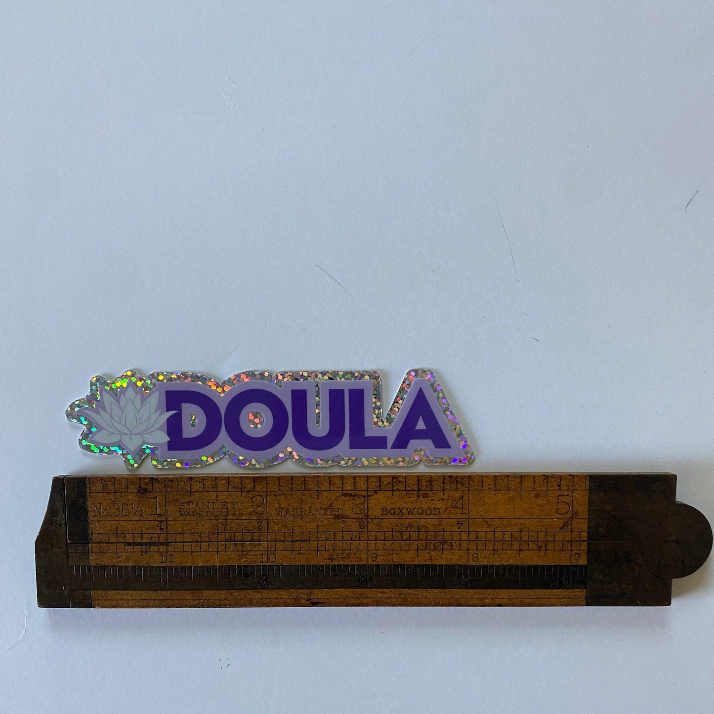 Glistening 'DOULA' sticker being measured by an antique wooden ruler on a white background.