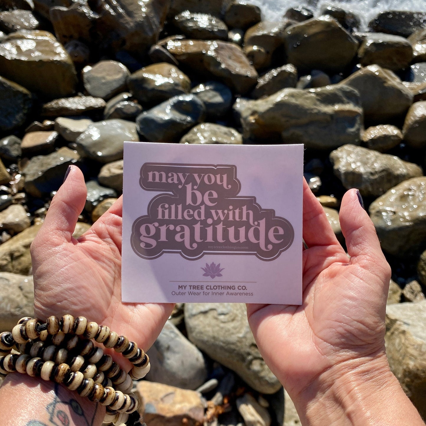 Hand holding a pink and brown sticker that reads, may you be filled with gratitude, against a rocky background.