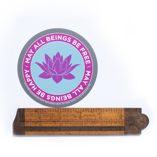 4" circular Buddhist sticker with a purple lotus flower and text, may all beings be free may all beings be happy, accompanied by a wooden ruler.