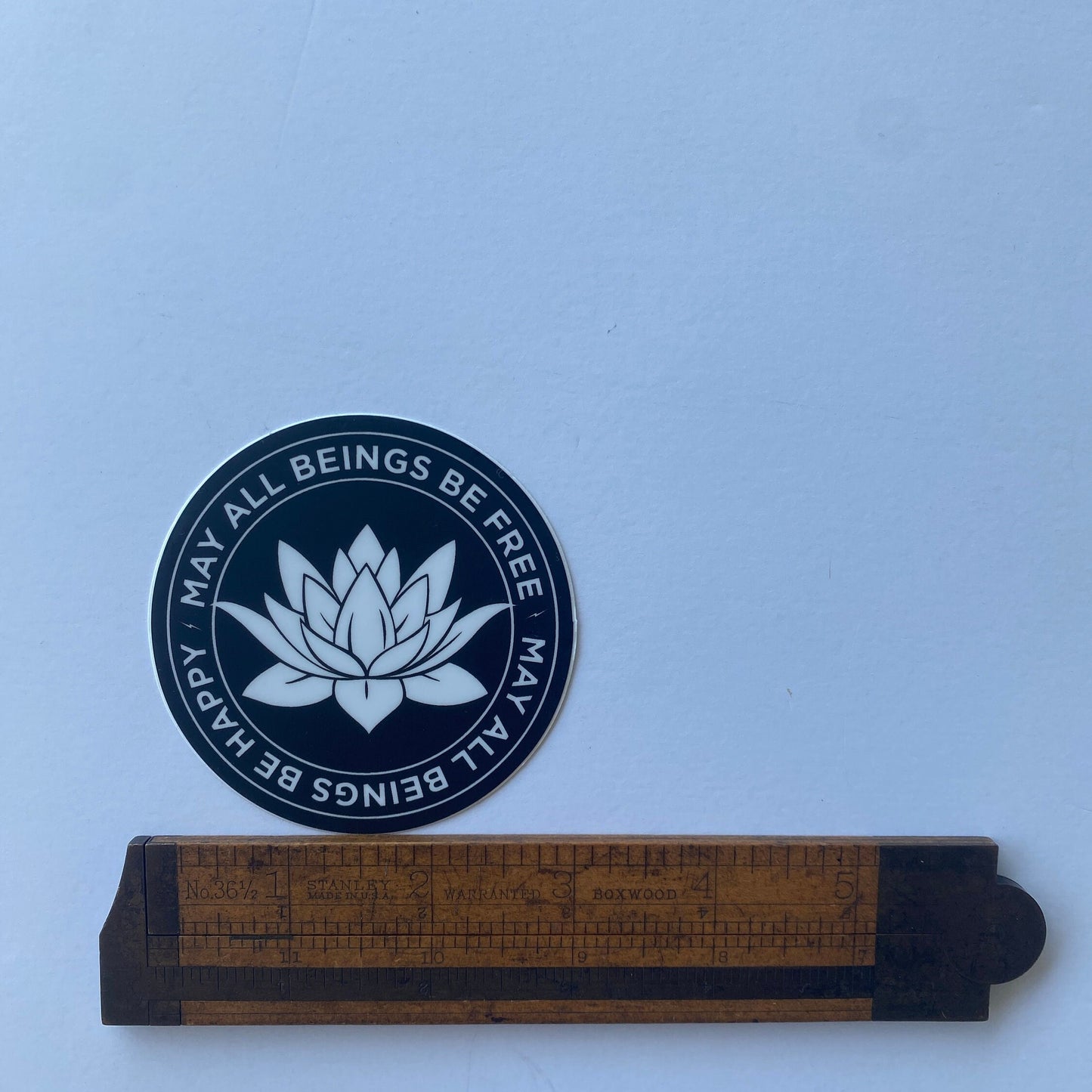3" circular sticker with a white lotus flower design and text, may all beings be free may all beings be happy, being measured by an antique wooden ruler.