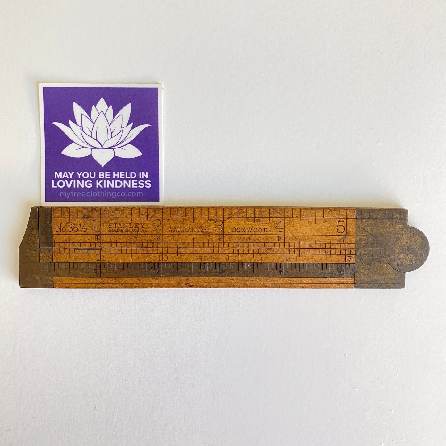 Wooden ruler with a 2" square purple and white lotus sticker with the words, may you be held in loving kindness.