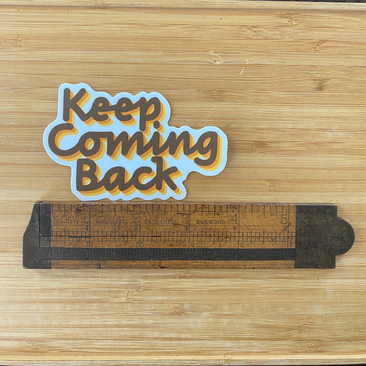 Decorative sign with 'Keep Coming Back' text on a wooden surface with a ruler for scale.