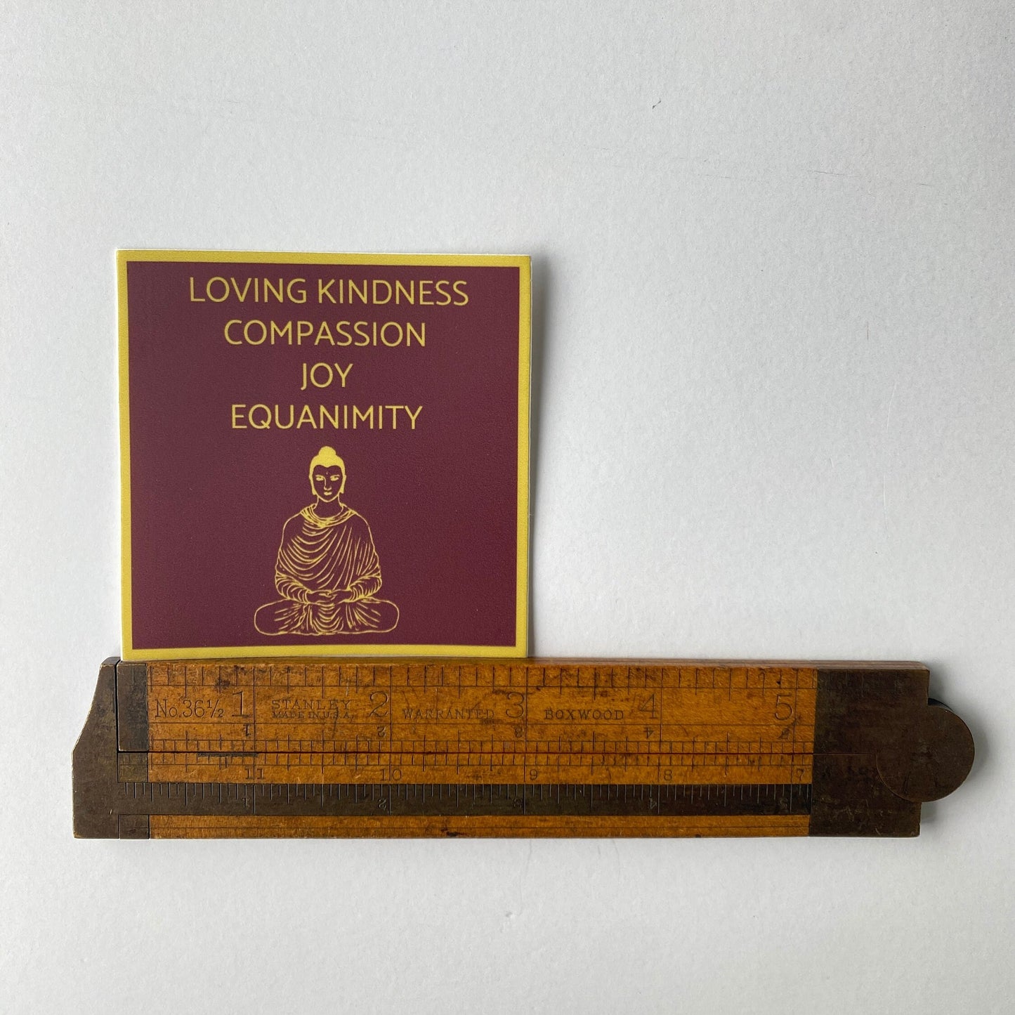 Three inch sticker with 'Loving Kindness, Compassion, Joy, Equanimity' text and a Buddha illustration on a white background, next to a wooden ruler.