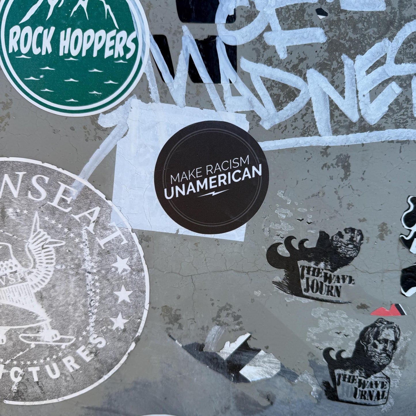 2.5" round black sticker with 'MAKE RACISM UNAMERICAN' on a concrete surface with various designs and text.