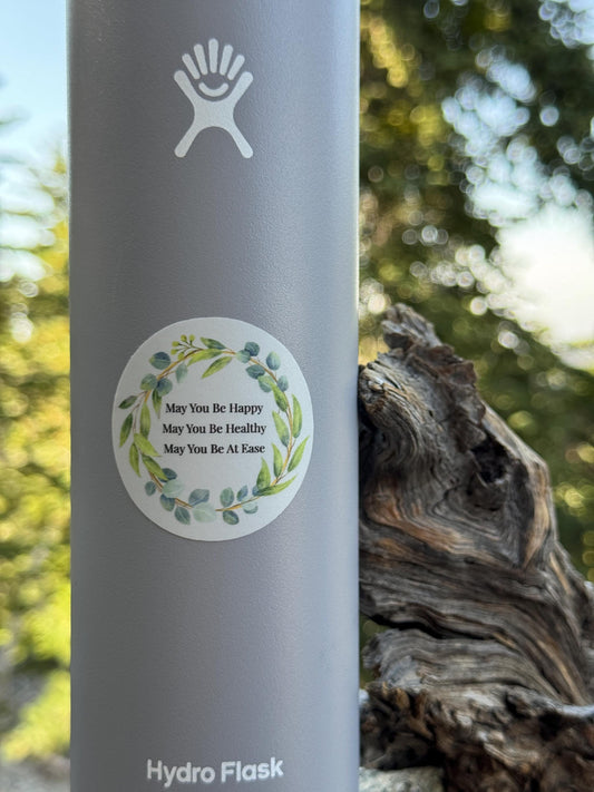 Hydro Flask water bottle with a 2" round blue sticker with Buddhist mettā prayer text, may you be happy may you be healthy may you be at ease, in a natural background.