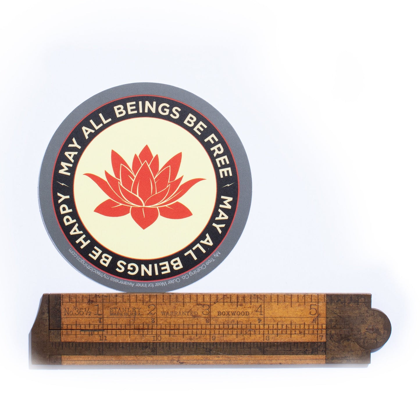 Round sticker with lotus flower design and text 'May all beings be free' on a white background, with a wooden ruler below.