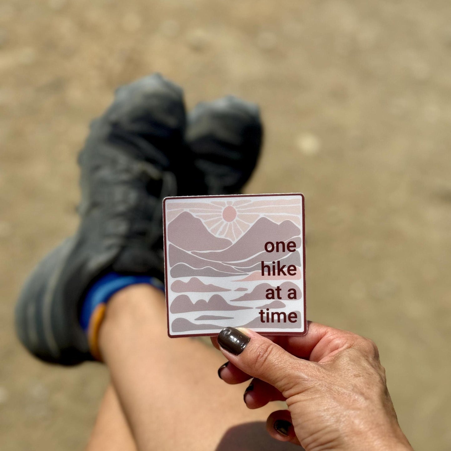 Hand holding a 3" square sticker with 'one hike at a time' in neutral colors with mountains and a sunset , with hiking boots in the background.