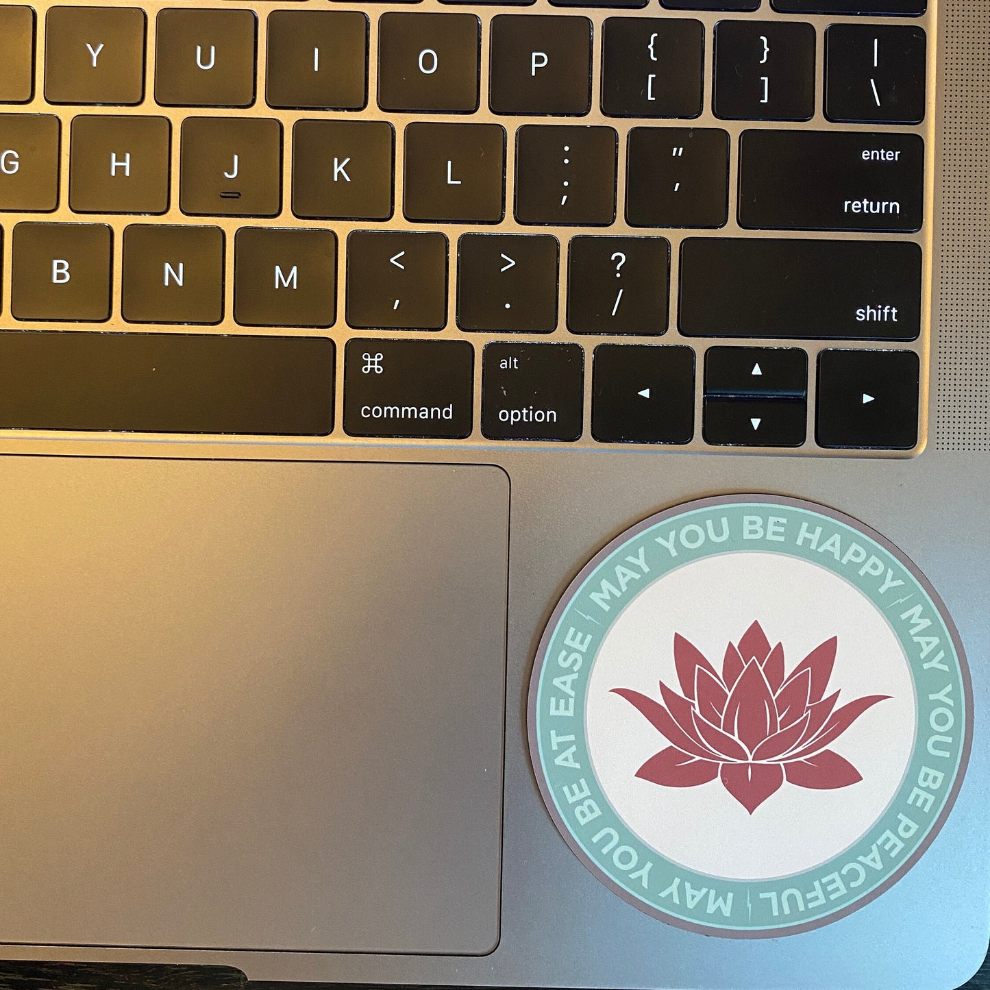 Laptop keyboard with a sticker featuring a lotus flower and text, "may you be happy may you be peaceful may you be at ease" on the palm rest area.