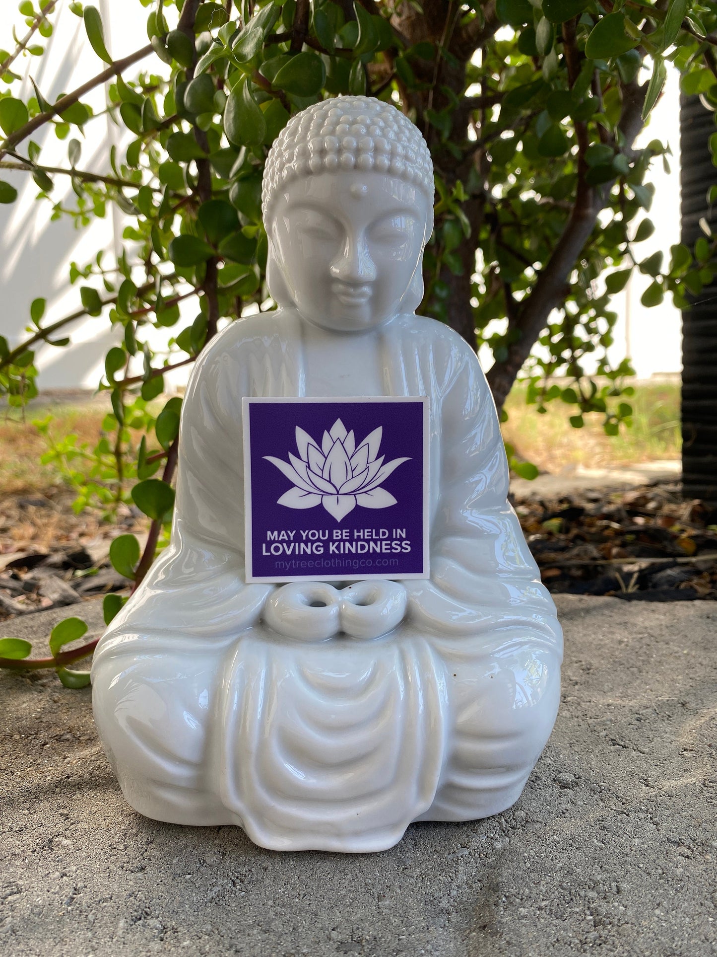 White Buddha statue with a 2" square purple and white lotus sticker with the words, may you be held in loving kindness.