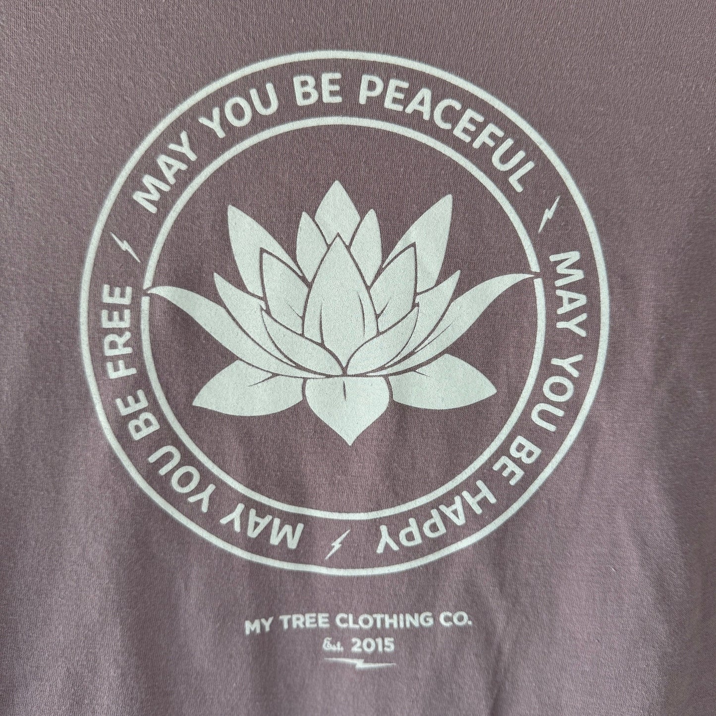 Close up photo of Buddhist shirt, light purple t-shirt with a white lotus in the center and the words, may you be peaceful may you be happy may you be free.