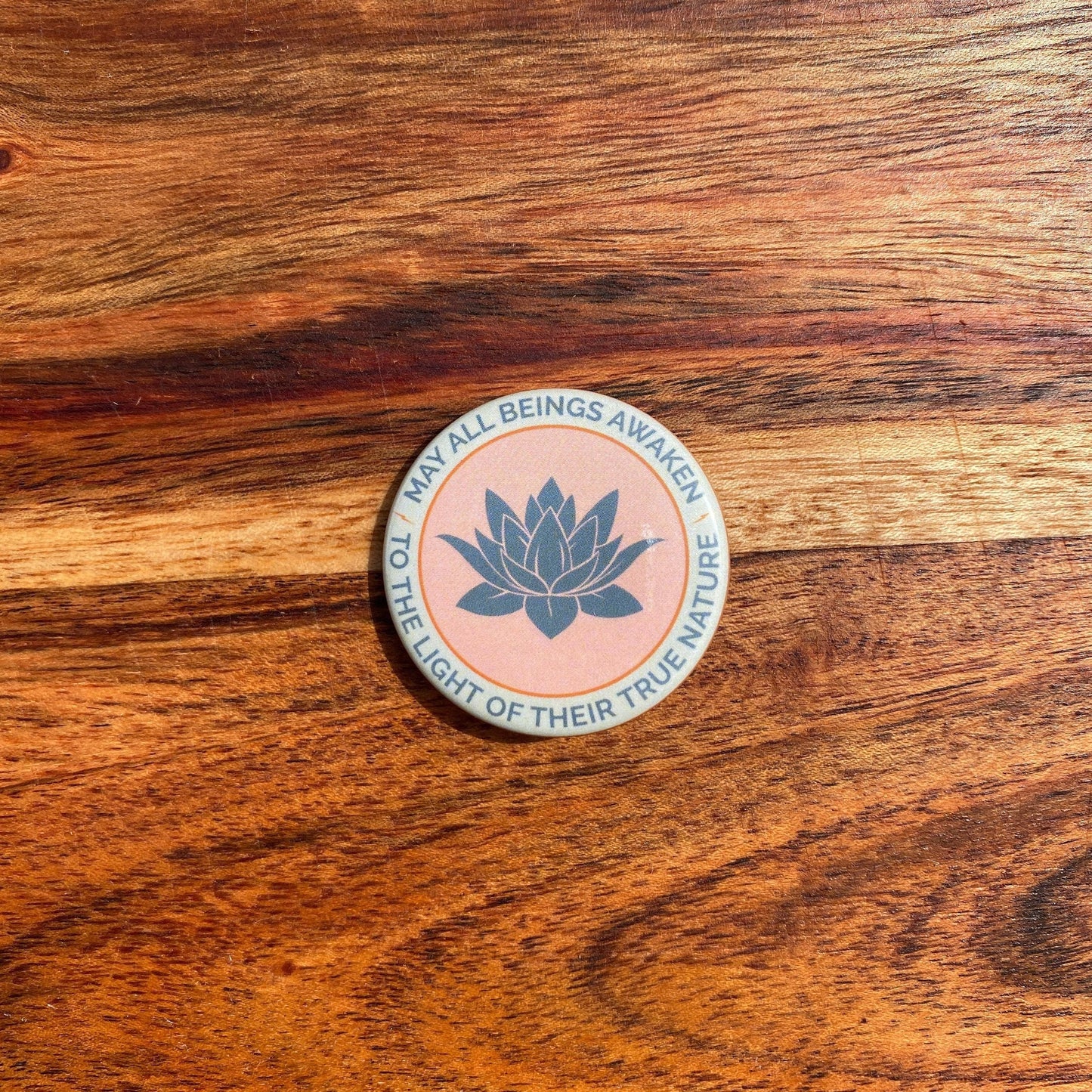 1.5" round Tibetan Buddhist lotus design button with the text, may all beings awaken to the light of their true nature, on a wooden background.