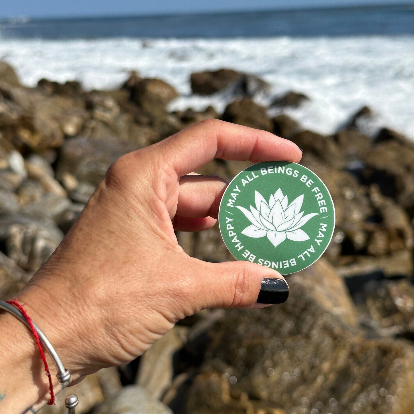 Hand holding a 2" round green sticker with a white lotus Buddhist design and the text, may all beings be free may all beings be happy, against a rocky beach background