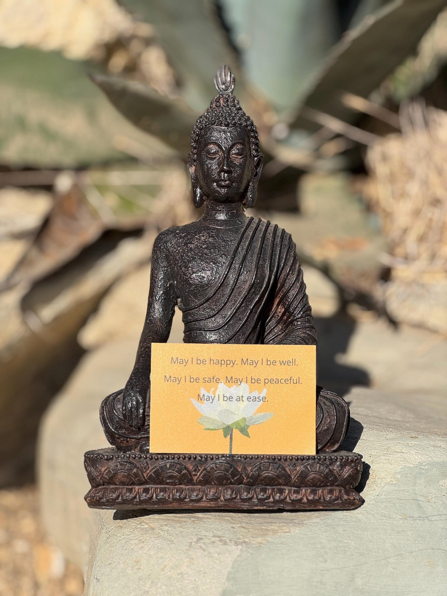 Buddha statue with a rectangular yellow sticker with a lotus and the words, may i be happy may i be well may i be safe may i be peaceful, on a rock with plants in the background.