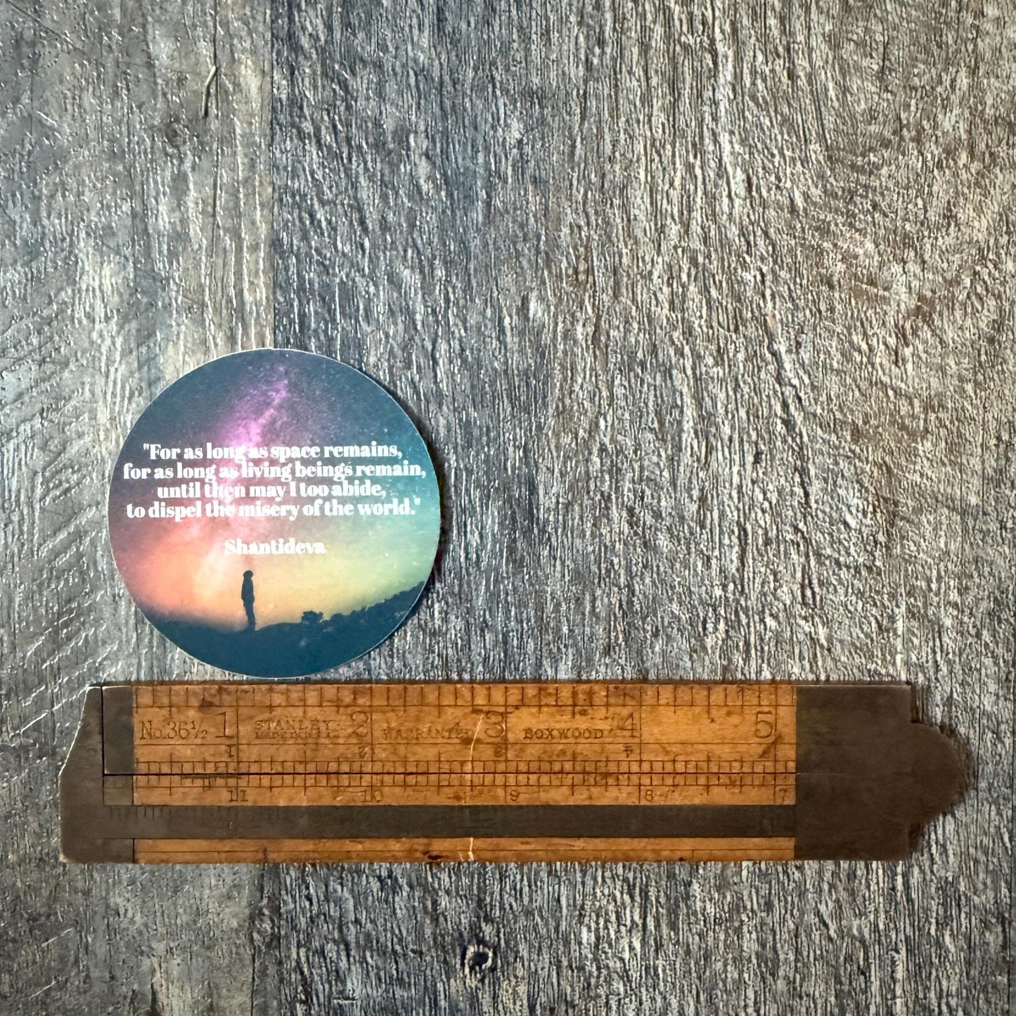 Round sticker with text and silhouette of a person standing on a hill, placed on a wooden surface with a ruler for scale.