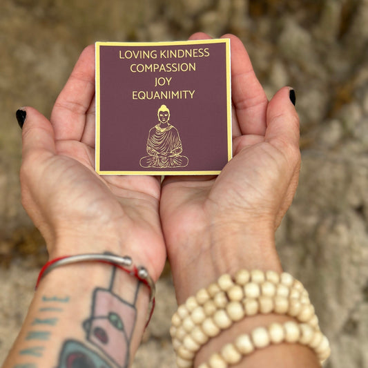 Hands holding a Tibetan red and gold three inch sticker with 'Loving Kindness, Compassion, Joy, Equanimity' text with a Buddha illustration and text on a natural background.