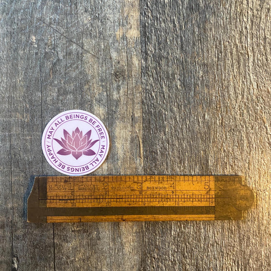 2" purple sticker with a lotus design and the text, may all beings be free may all beings be happy on a wooden ruler against a wooden background.