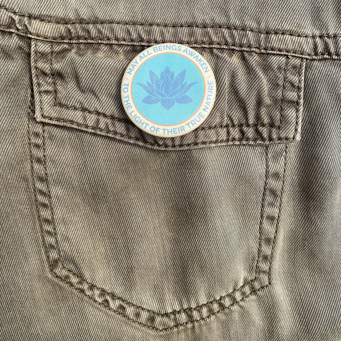 1.5" button with the mantra that reads, may all beings awaken to the light of their true nature, with a lotus design on a gray denim pocket.
