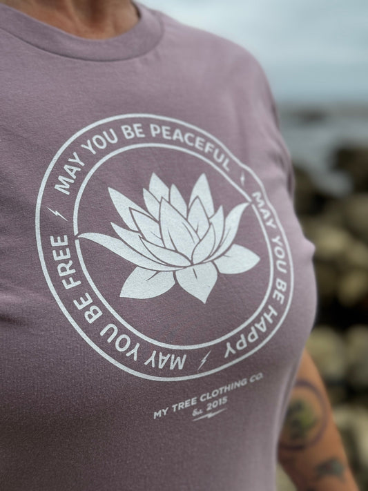 Close up of person wearing a purple t-shirt with a white lotus flower design and text on a blurred natural background.