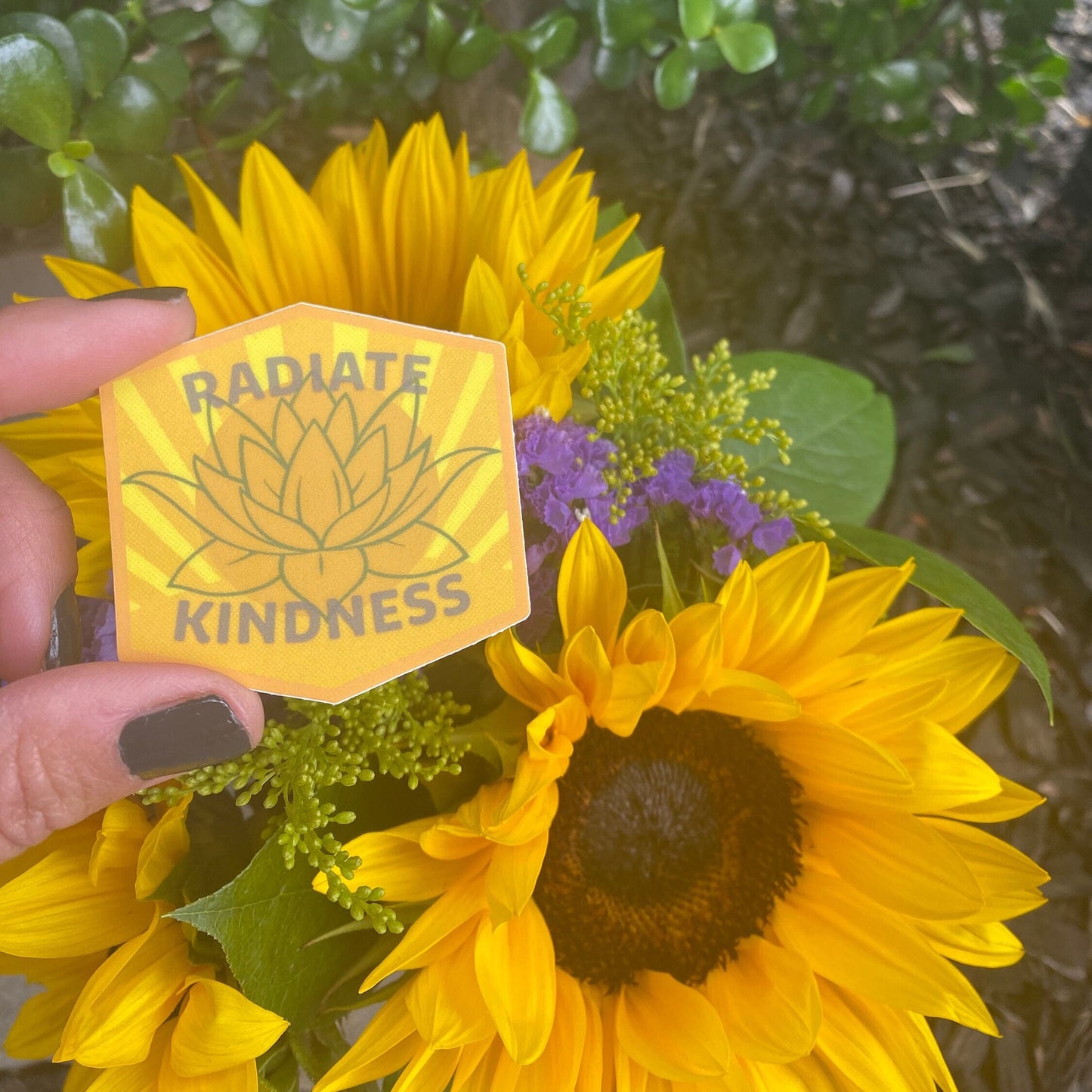 Hand holding a 'Radiate Kindness' sticker among sunflowers and greenery